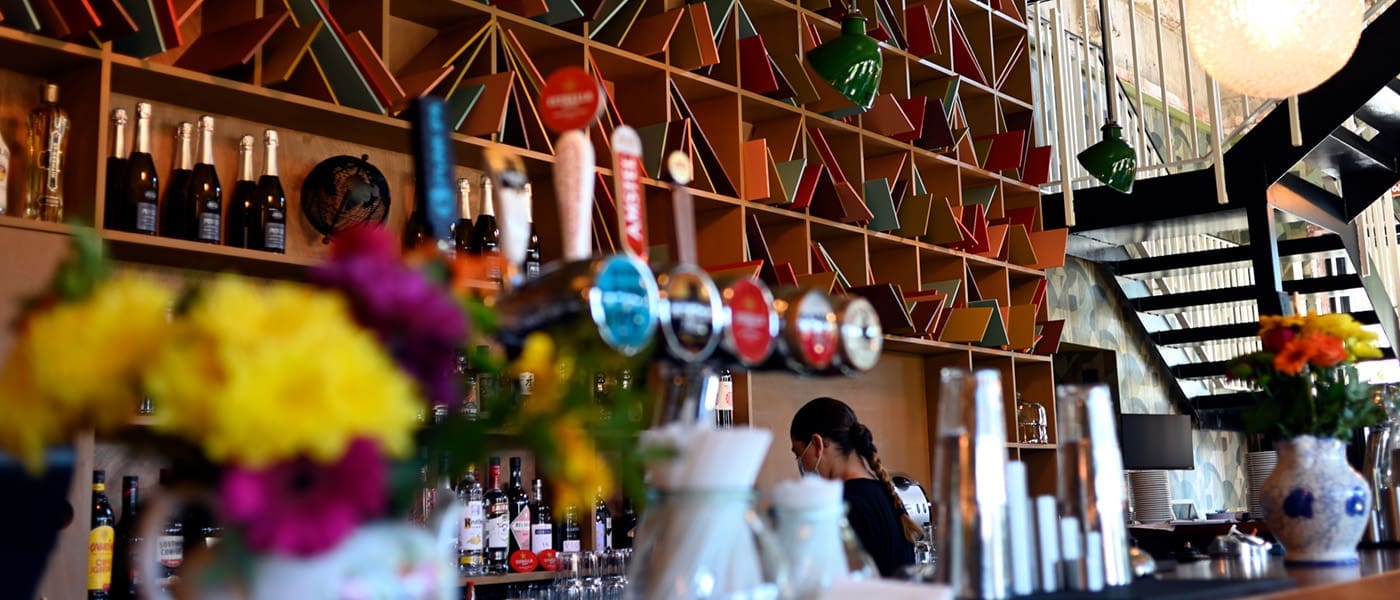 A colorful Postino bar with geometric shelves holding bottles and decor, fresh flowers on the counter, beer taps in the foreground, and two people behind the bar under modern lighting near a staircase.