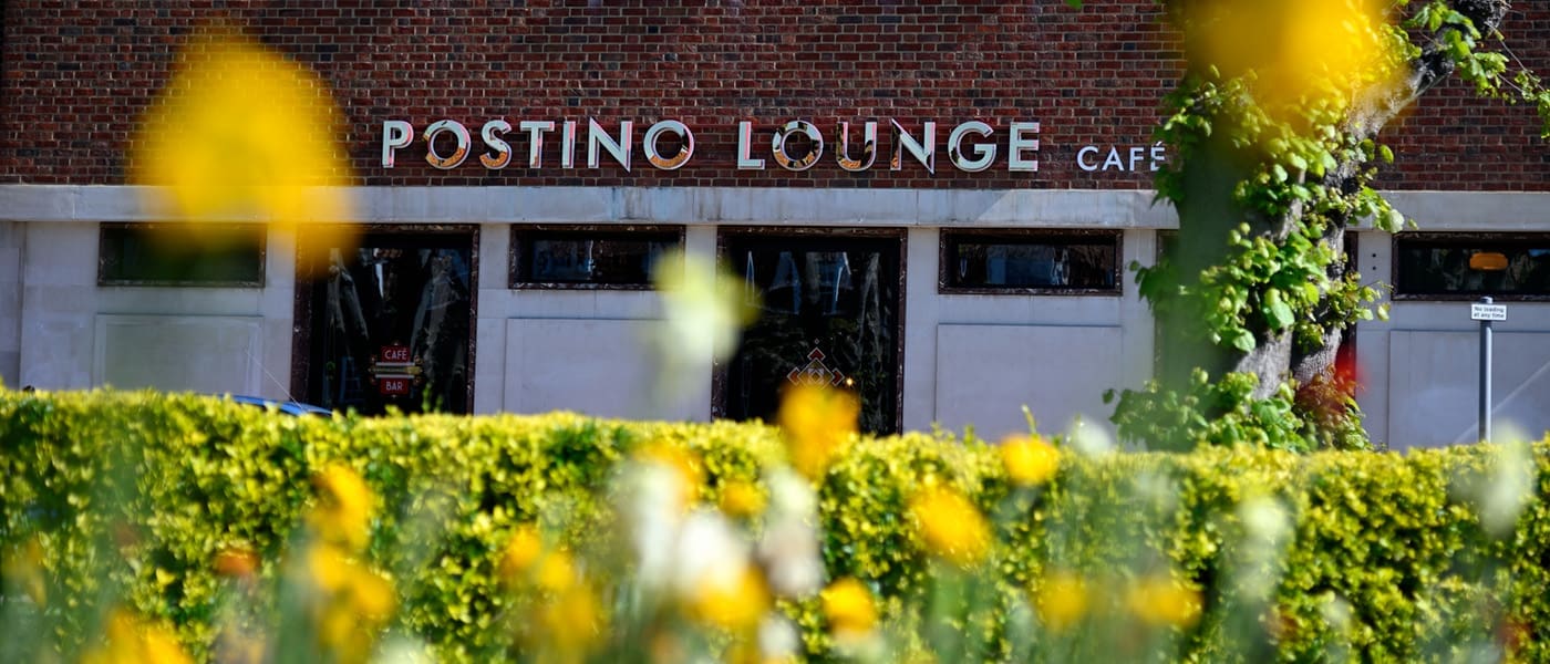 The exterior of Postino Lounge Café, with its Postino sign visible on a brick building behind a green hedge and blurred yellow flowers in the foreground.
