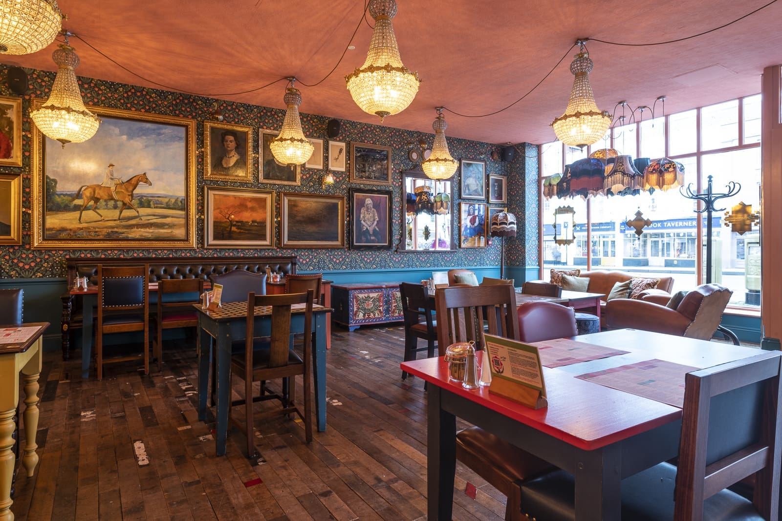 A cozy, eclectic café named Prospero with wooden floors, mismatched tables and chairs, chandeliers, and colorful walls decorated with numerous framed paintings and portraits. Large windows let in natural light.
