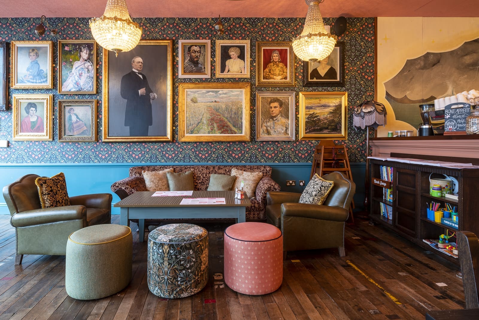 A cozy, eclectic lounge with vintage armchairs, ottomans, and a Prospero-patterned sofa beneath a wall of framed portraits and landscapes. Chandeliers hang from the ceiling, and art supplies fill a dark wood shelf on the right.