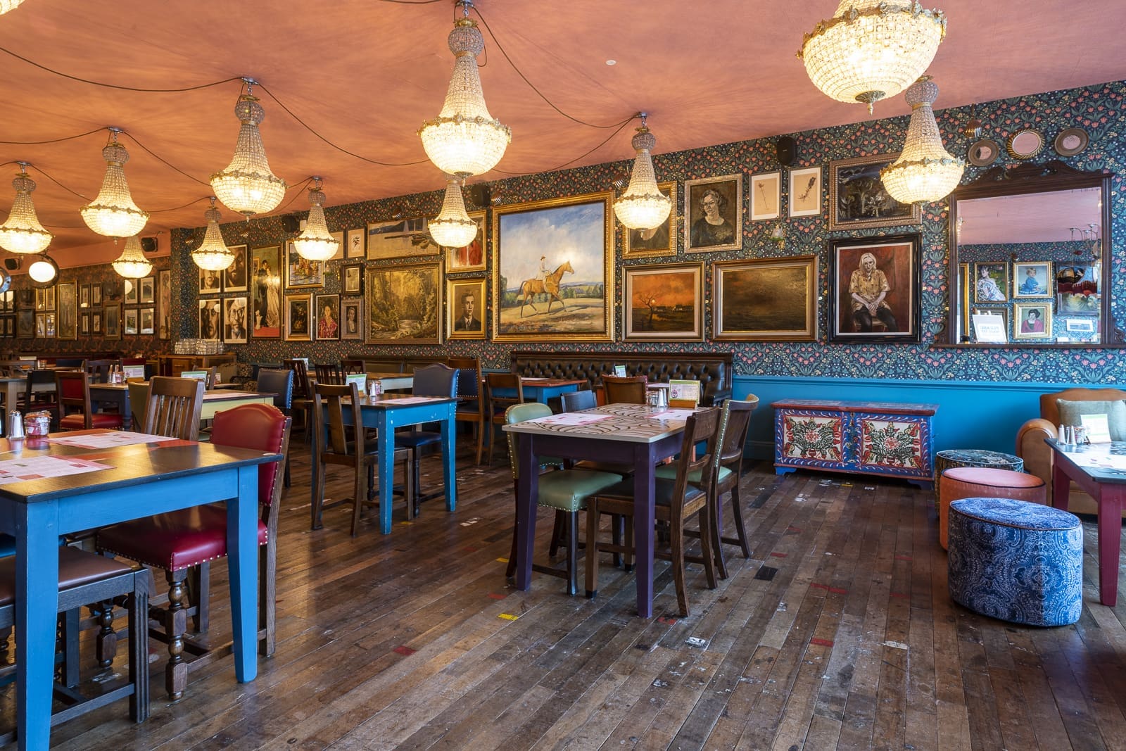 Eclectic restaurant interior with colorful mismatched tables and chairs, ornate chandeliers, wood flooring, and walls covered in framed paintings and art creates a cozy, artistic atmosphere reminiscent of Prospero’s vibrant imagination.