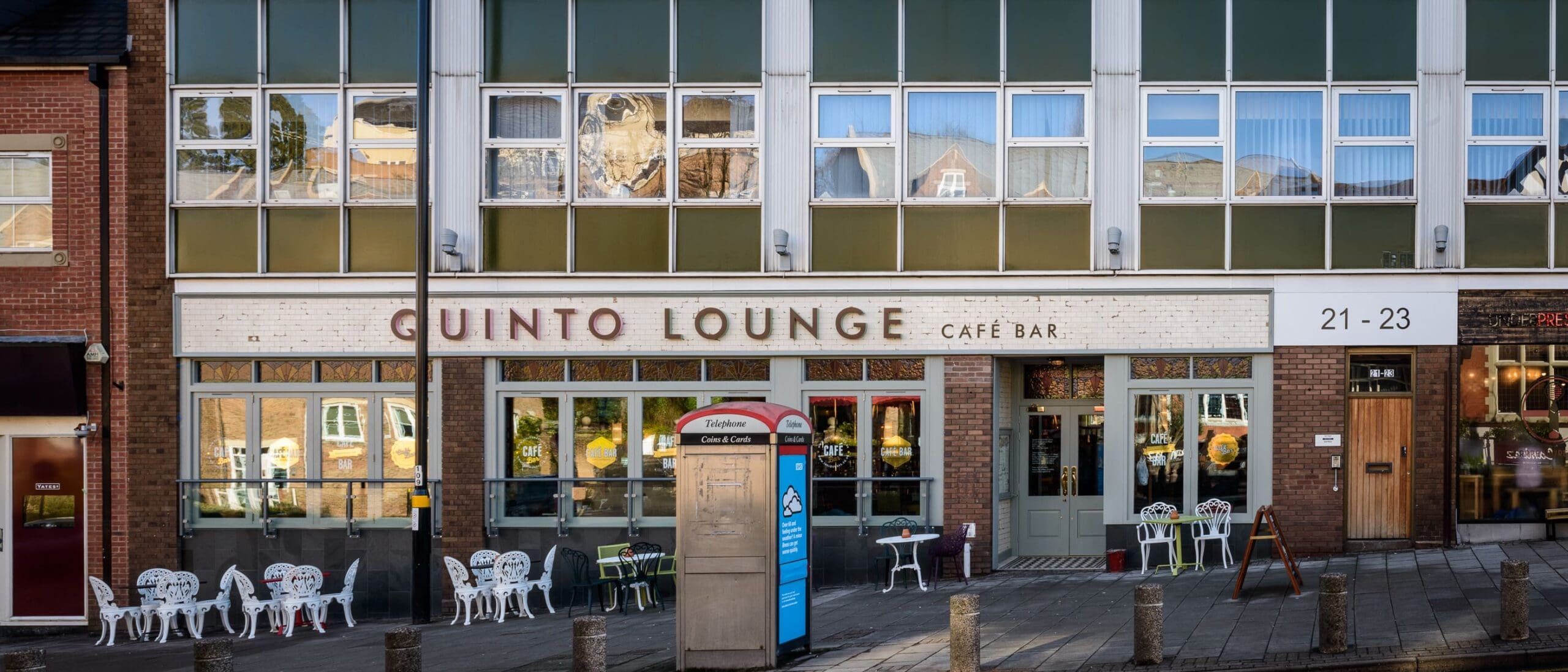 Quinto Lounge café bar exterior with large windows, a classic red telephone booth, white outdoor chairs and tables on the pavement, and building number 21-23 visible on the right. The inviting Quinto atmosphere welcomes all.