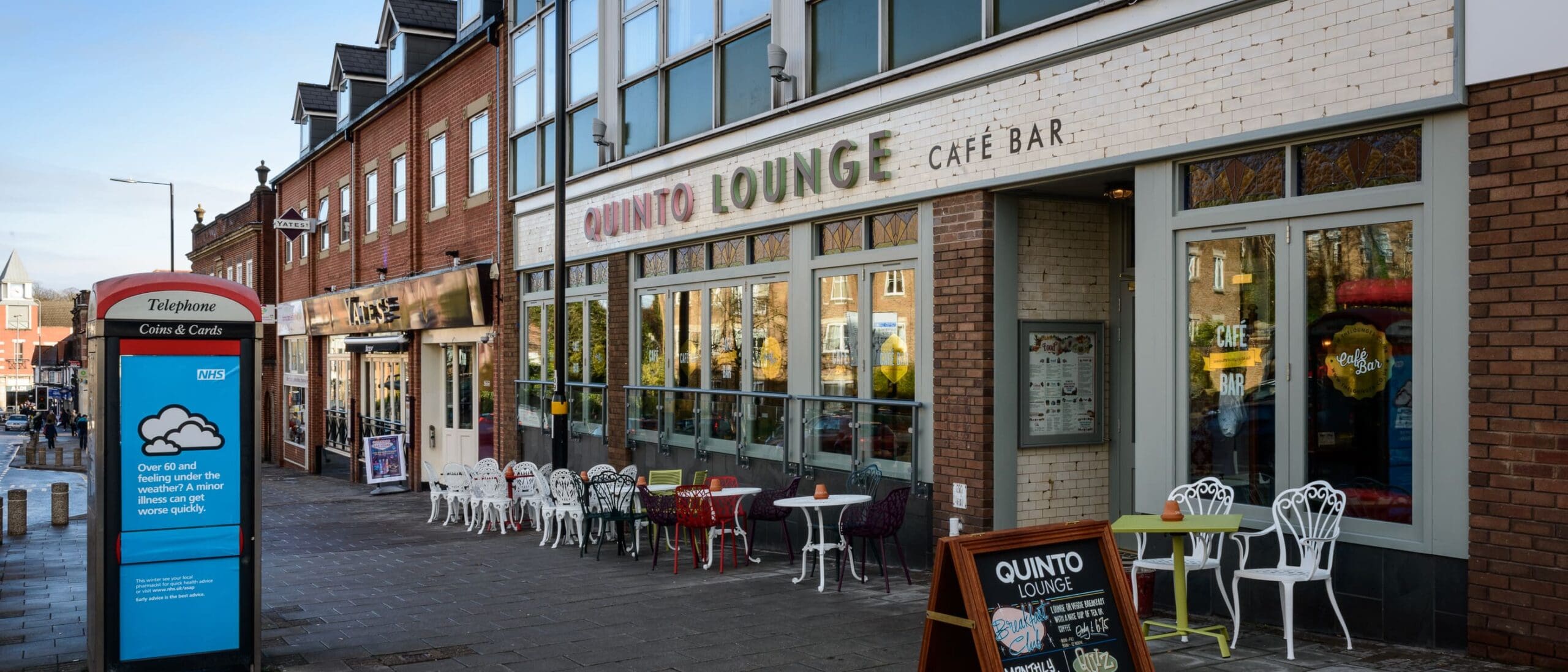 Street view of Quinto Lounge café bar with outdoor seating, colorful chairs and tables, a menu board, and a red British telephone box nearby on the pedestrian sidewalk outside Quinto.