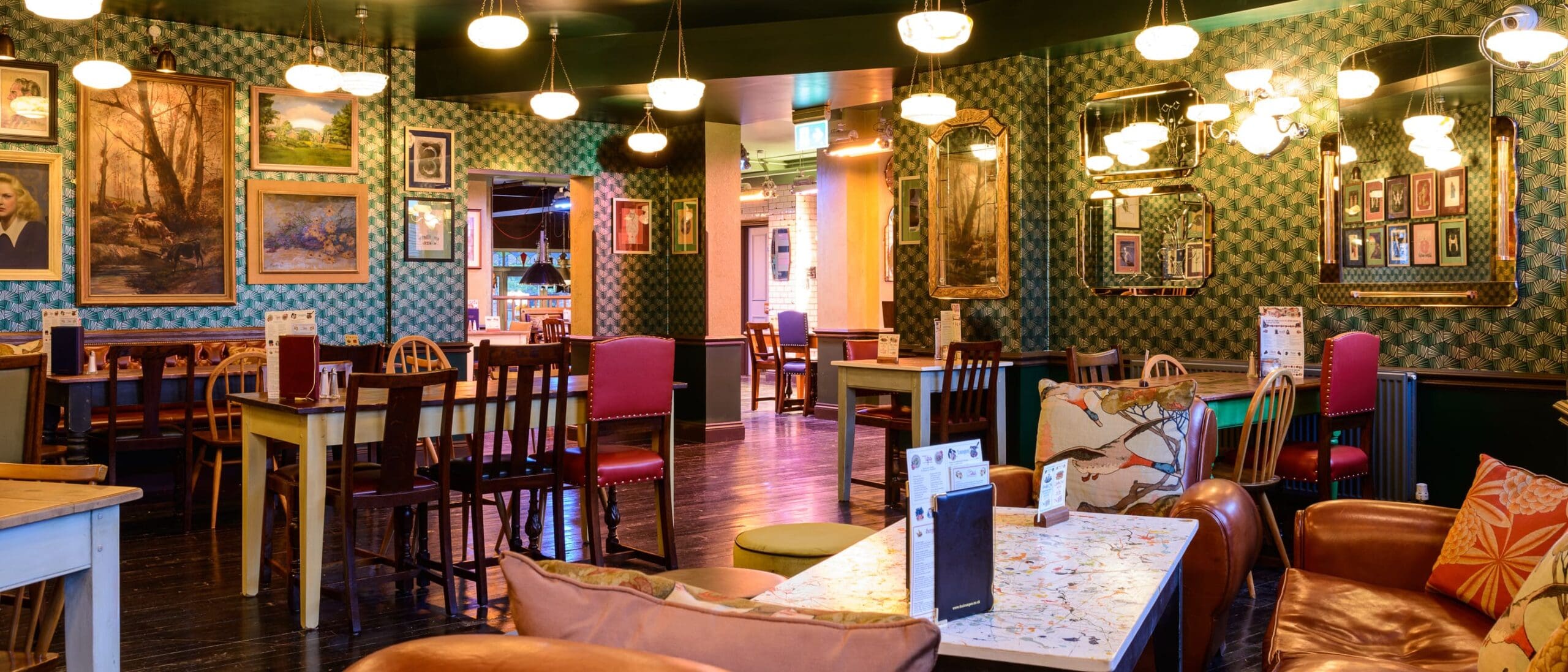 A cozy, eclectic cafe in Quinto with patterned wallpaper, framed art on the walls, mismatched chairs, cushioned armchairs, and tables set for dining under warm hanging lights.
