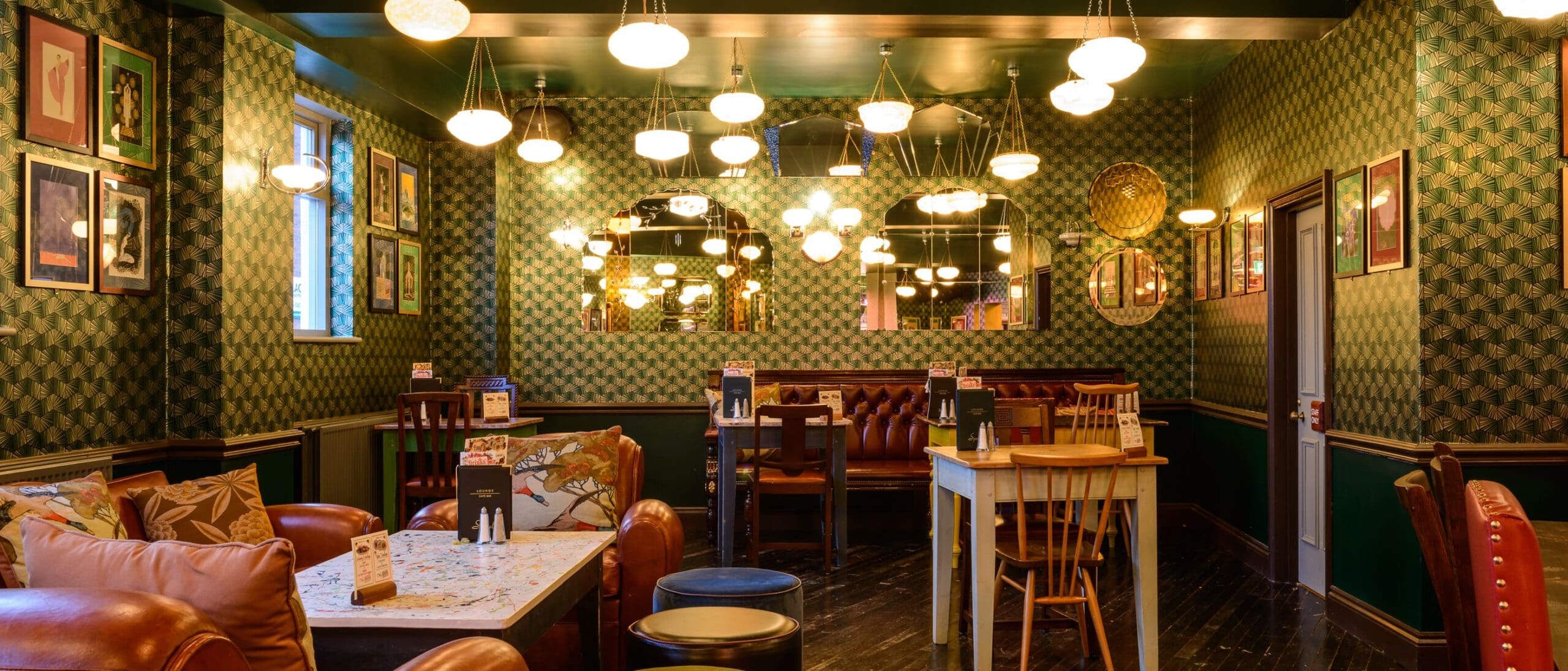 A cozy, stylish pub interior with patterned wallpaper, hanging lights, cushioned seating, framed art on the walls, wooden tables, and a mix of chairs and benches creates Quinto’s warm, inviting atmosphere.