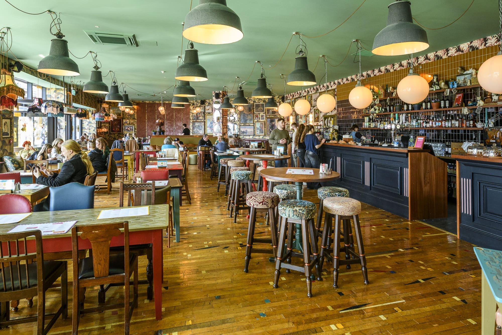 A cozy, colorful café with mismatched tables and chairs, pendant lights hanging from the ceiling, people chatting and relaxing in this inviting riparo, and a lively bar area where staff are serving drinks.