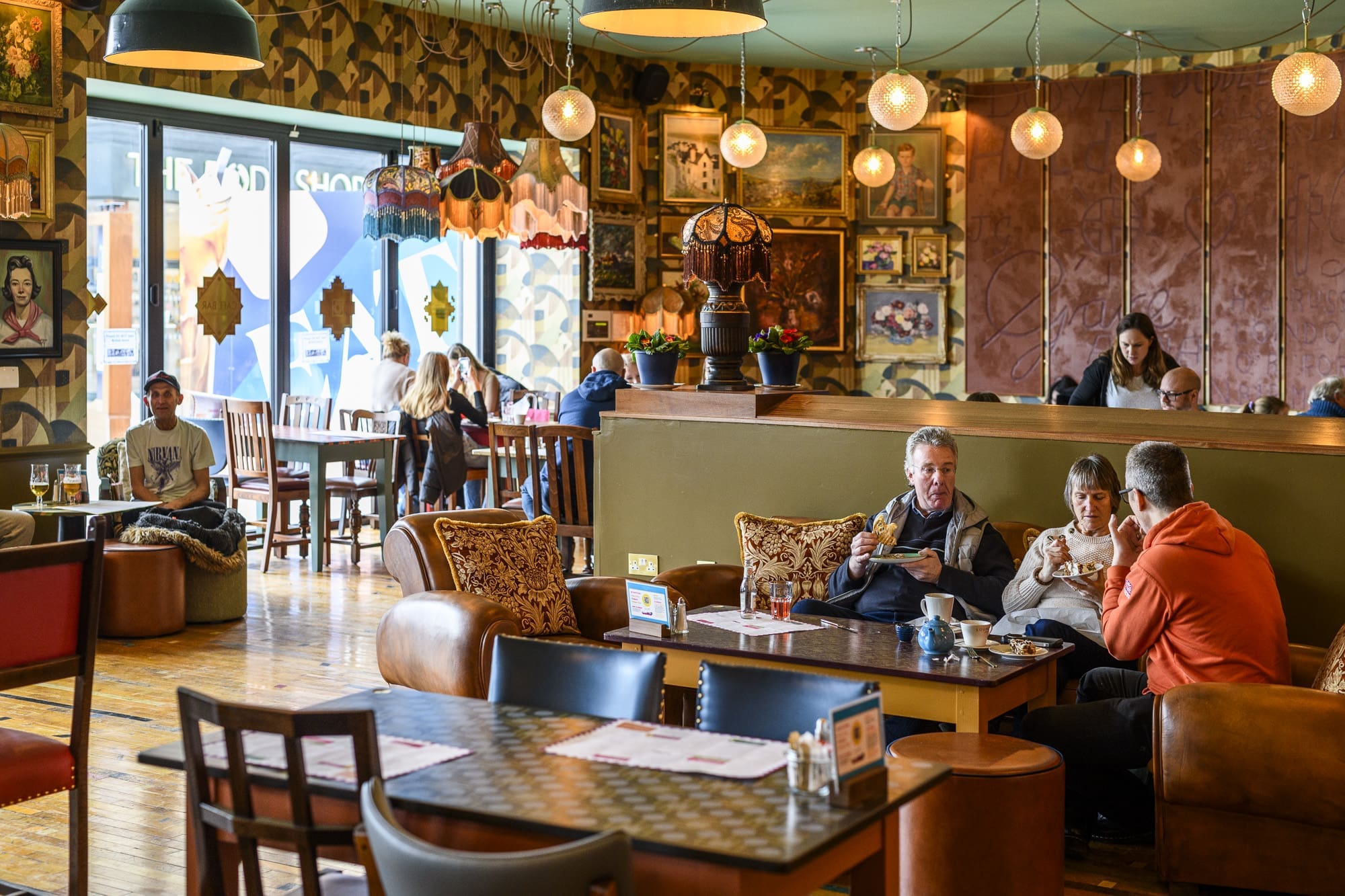 A cozy, eclectic café with vintage decor, warm lighting, and people enjoying food and conversation. The walls are decorated with paintings and lamps, while large windows let in natural light—a charming riparo from the bustle outside.