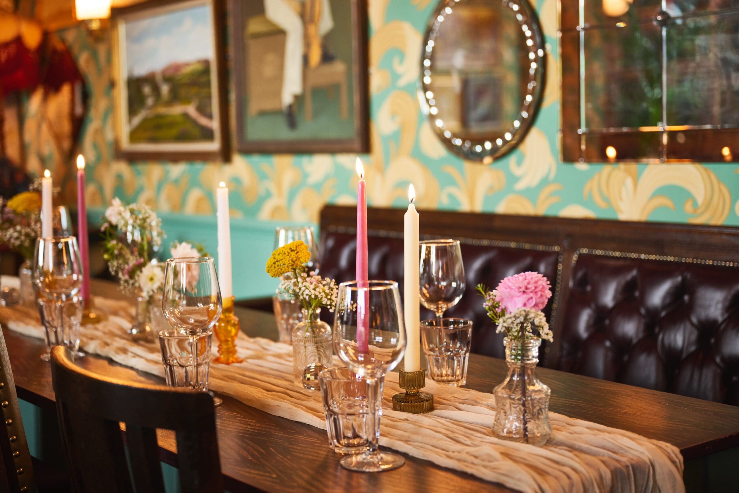 A wooden table set for a meal with candles, small vases of flowers, glassware, and napkins. The background features patterned teal walls, framed art, mirrors, and a tufted leather bench.