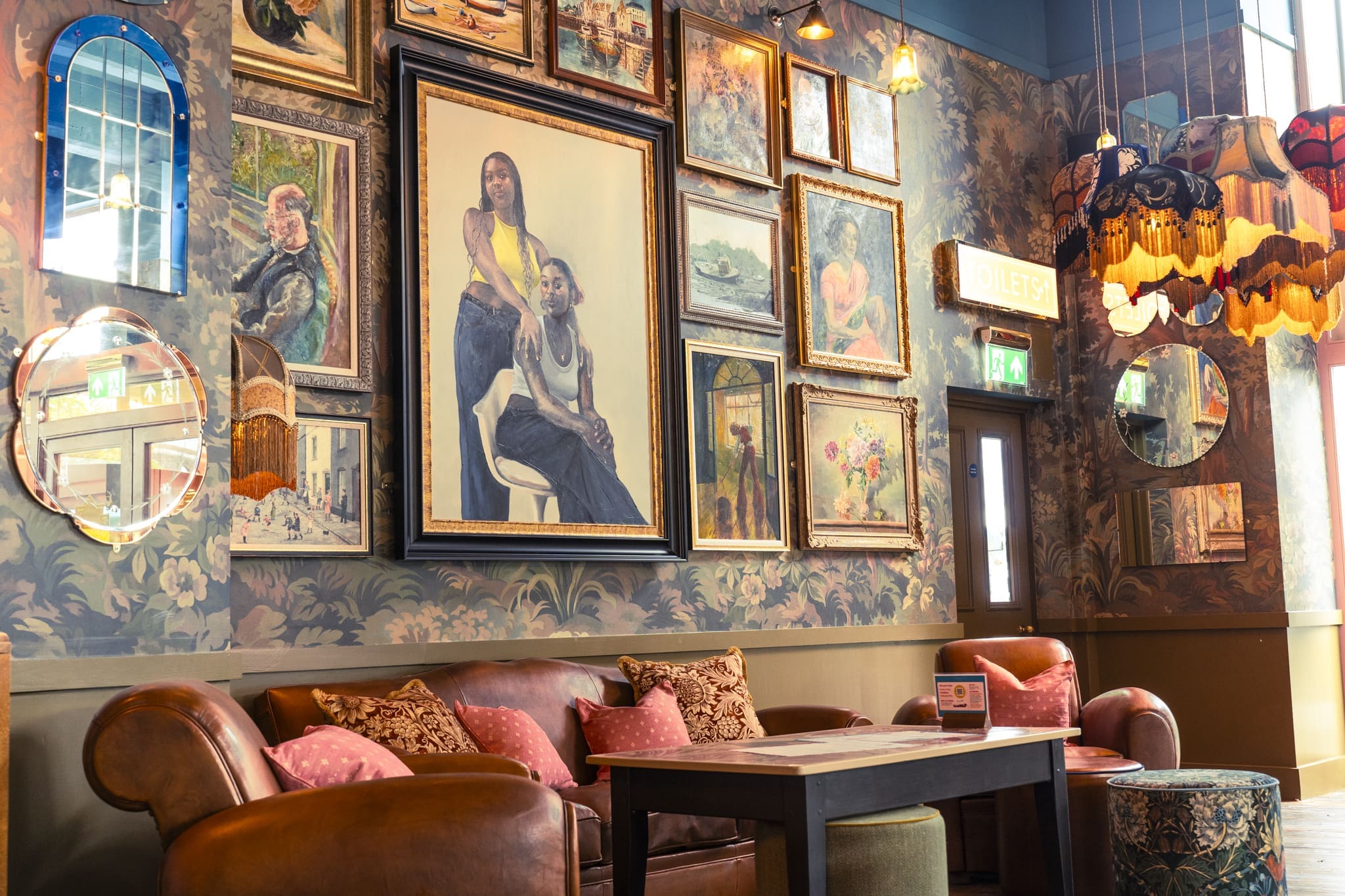 A cozy vintage-style lounge, Ritorno, with leather sofas, patterned wallpaper, and a wall filled with framed artwork and paintings. Warm lamps and mirrors add to the eclectic, inviting atmosphere.
