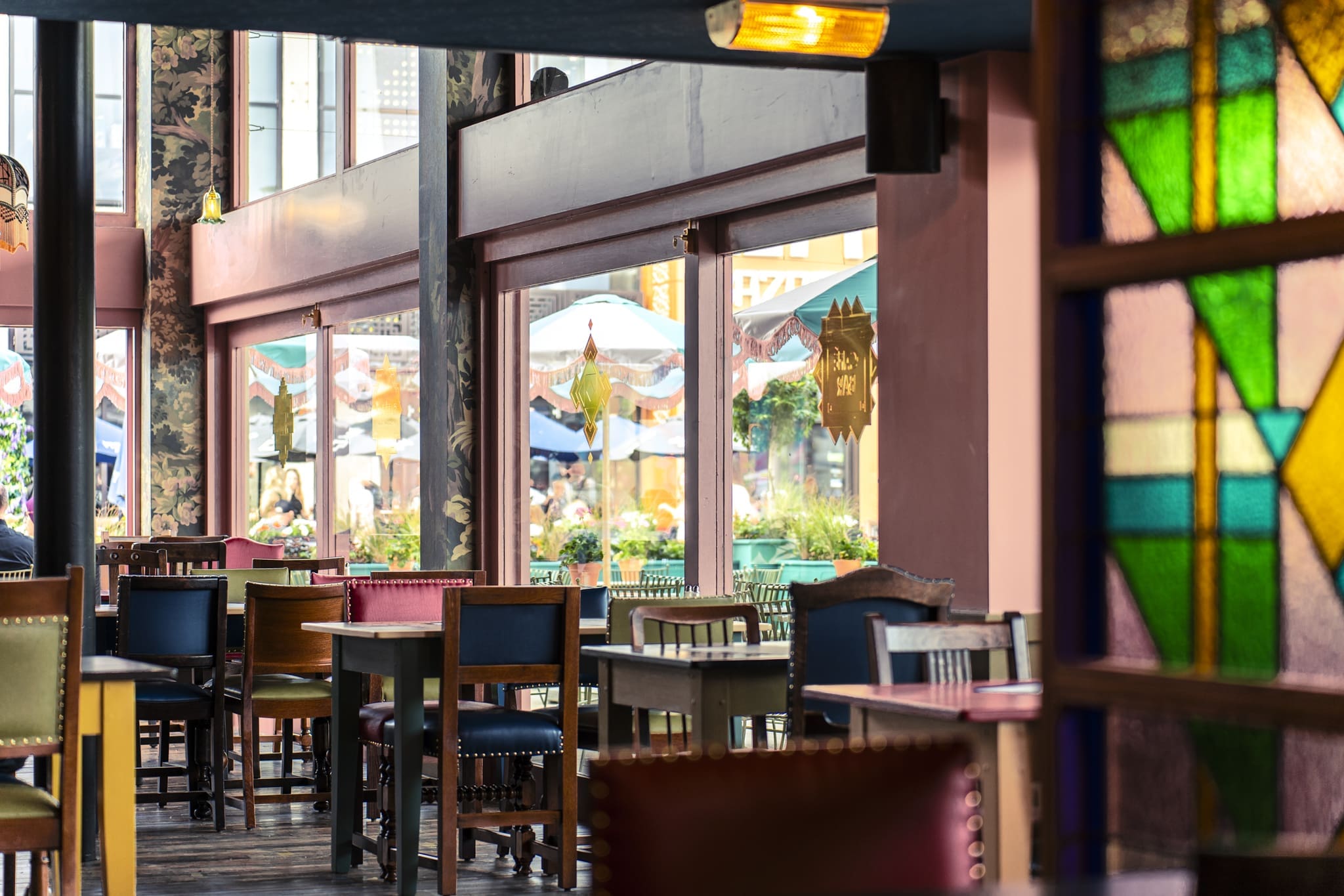 A colorful, eclectic restaurant interior at Ritorno features mismatched chairs and tables, large windows, floral columns, and stained glass panels. Outdoor seating with umbrellas and greenery is visible through the windows.