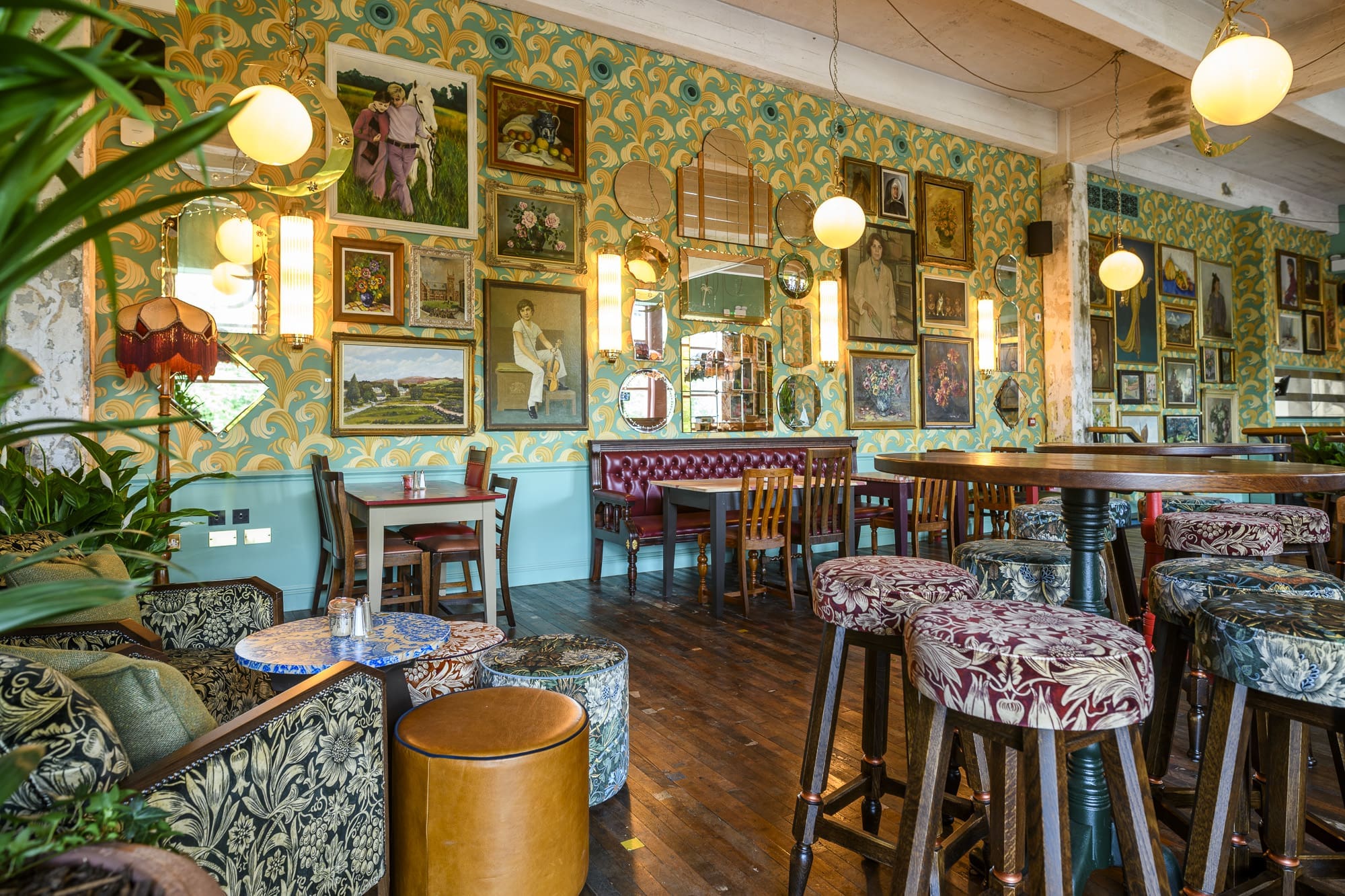 Eclectic bar interior with colorful patterned wallpaper, vintage paintings and mirrors on the walls, assorted wooden tables and chairs, floral upholstered stools, and warm hanging globe lights, creating a cozy, artistic atmosphere.