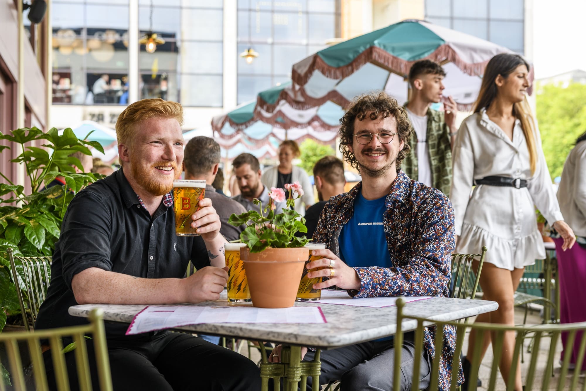 Two smiling men sit at an outdoor cafe table with Ritorno beers, surrounded by plants and people. Other patrons and colorful umbrellas add to the lively background.