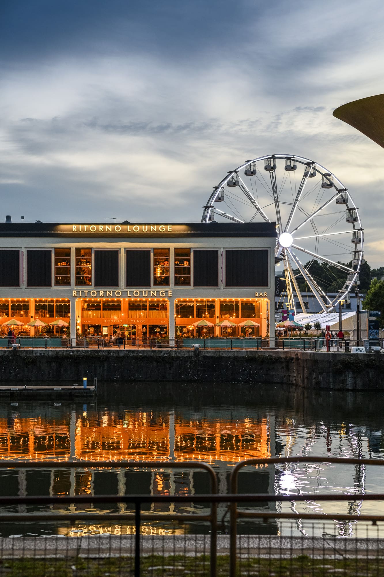 A modern building labeled "Ritorno Lounge" with glowing lights reflects on a waterfront at dusk, with a large illuminated Ferris wheel and partly cloudy sky in the background.