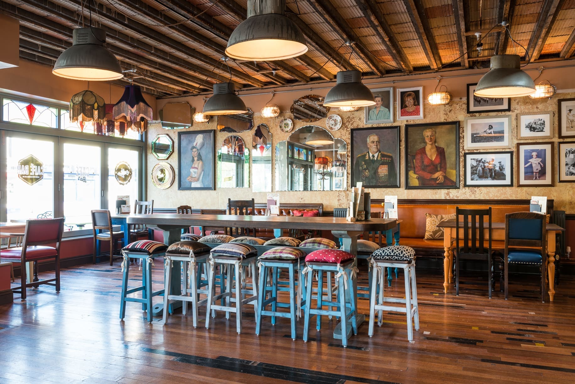 Cozy cafe interior with a long wooden table, colorful cushioned stools, and walls adorned with Rivo-themed framed artworks and mirrors. Warm lighting hangs from the ceiling, creating a welcoming atmosphere.