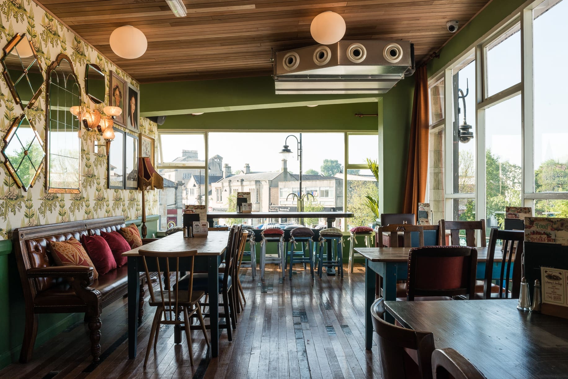 A cozy, sunlit café with wooden floors, vintage chairs, patterned wallpaper, and large windows offering views of buildings and greenery outside—Rivo’s inviting décor includes decorative mirrors, colorful cushions lining a bench, and tables set for dining.