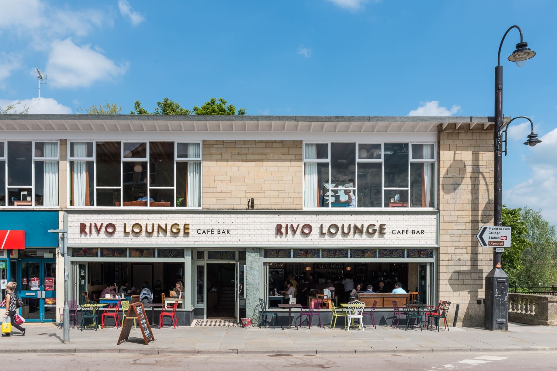 A street view of Rivo Lounge Café Bar, with outdoor seating and people dining under a clear blue sky. The Rivo building features large windows and a sign above the entrance, while a street lamp and road sign stand on the right.