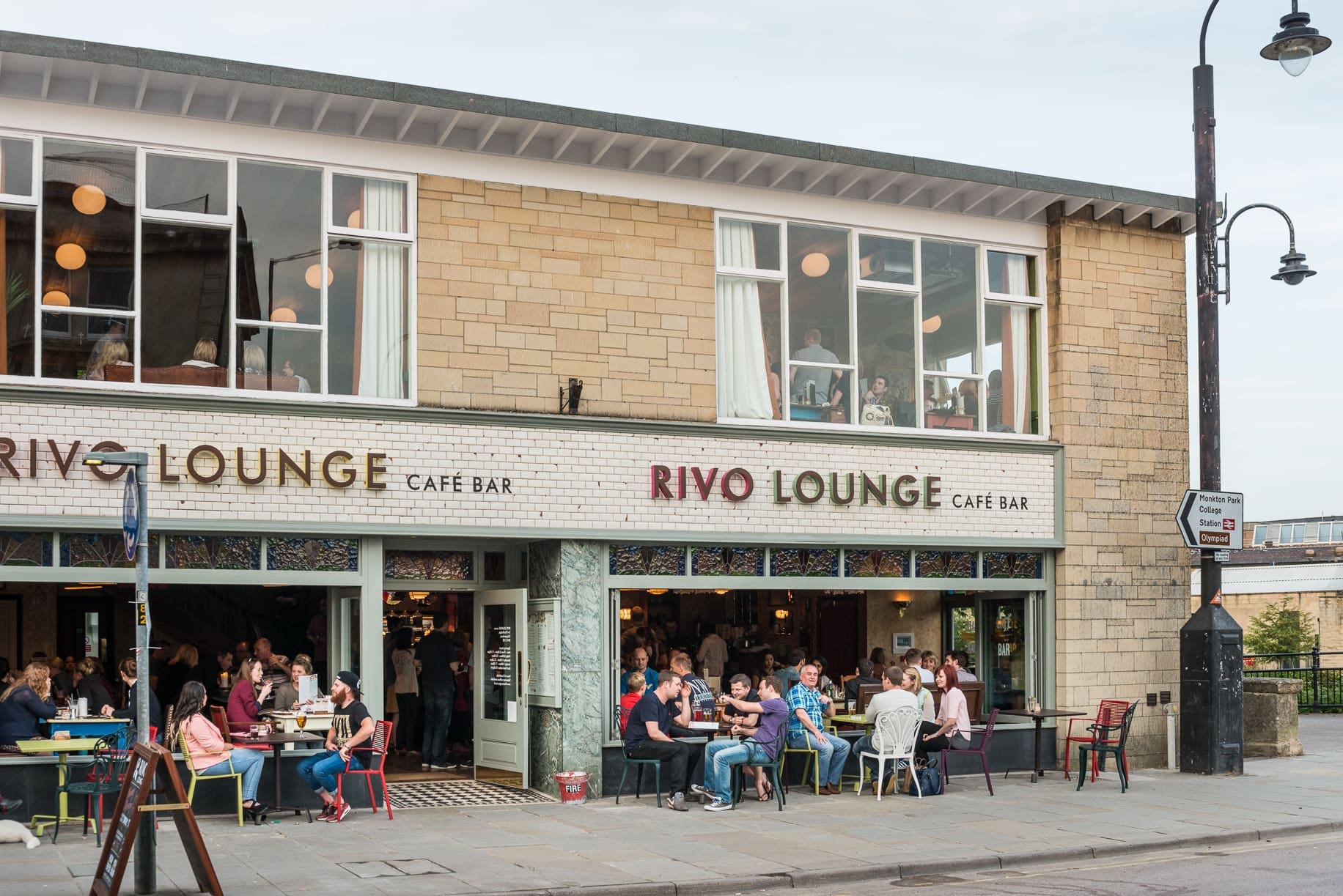 People are sitting outside and inside Rivo, a lively two-story café bar with large windows. Some enjoy eating and chatting at Rivo's sidewalk tables, while others relax indoors or upstairs.