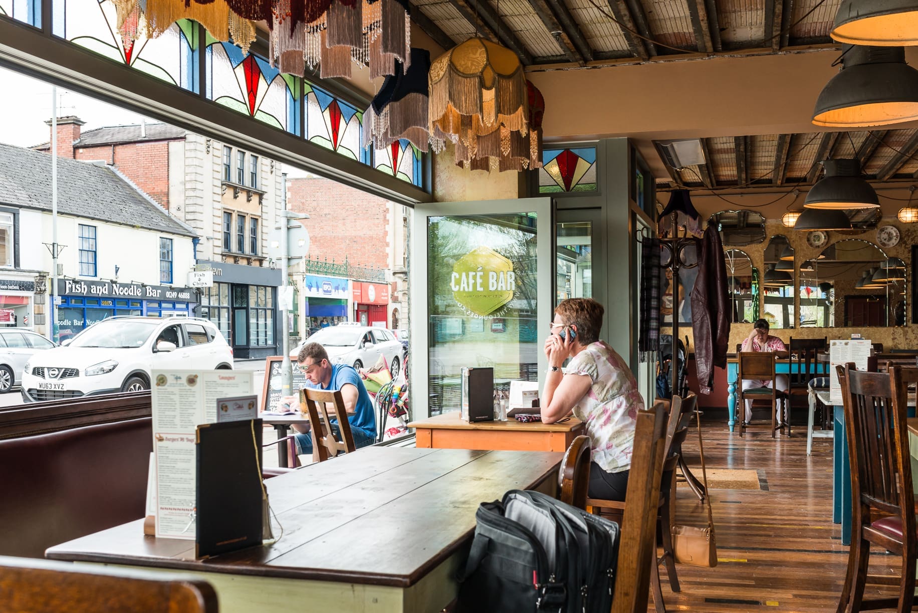 A cozy café bar, Rivo, with stained glass windows, wooden tables, and eclectic lampshades. A woman sits inside talking on the phone, while another person relaxes outside. Cars and shops are visible through the large front window.