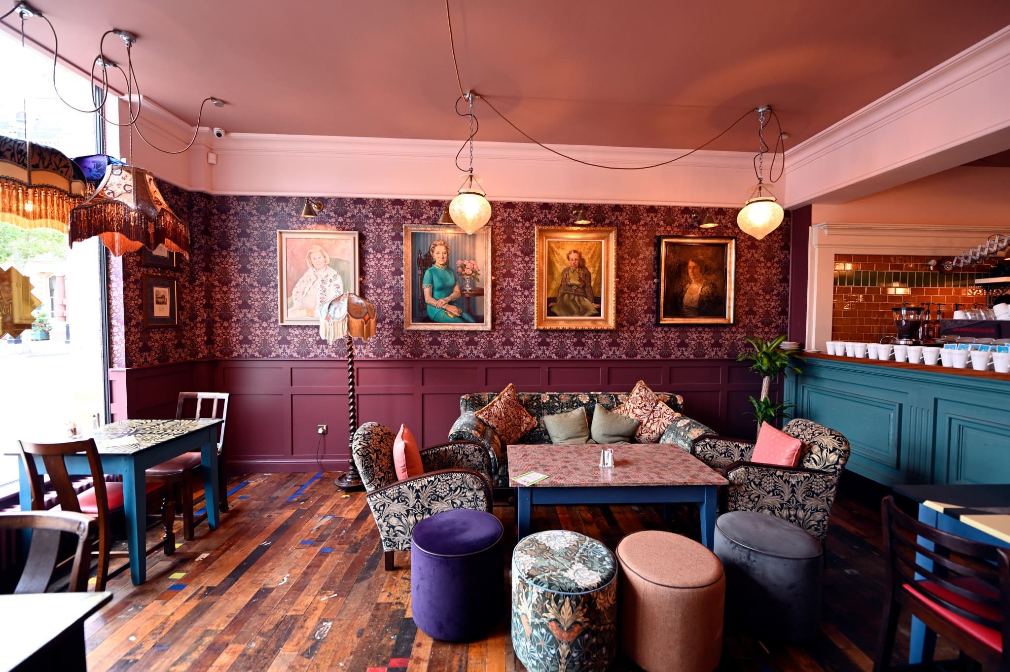 A cozy vintage-style cafe with patterned wallpaper, eclectic armchairs, mismatched tables and chairs, colorful cushions, and Rondo hanging lamps. Framed portraits adorn the walls, with wooden floors and a bar visible in the background.