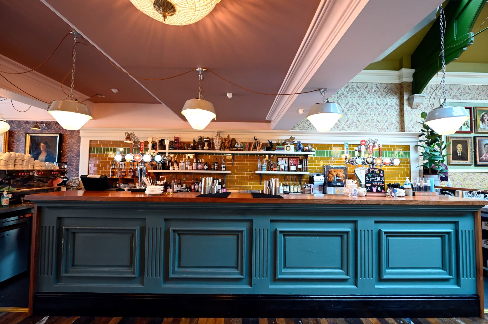 A stylish, empty bar with a turquoise counter and Rondo-inspired hanging white lamps. Shelves stocked with bottles and glassware line the walls, set against yellow tiles, framed art, and patterned wallpaper.