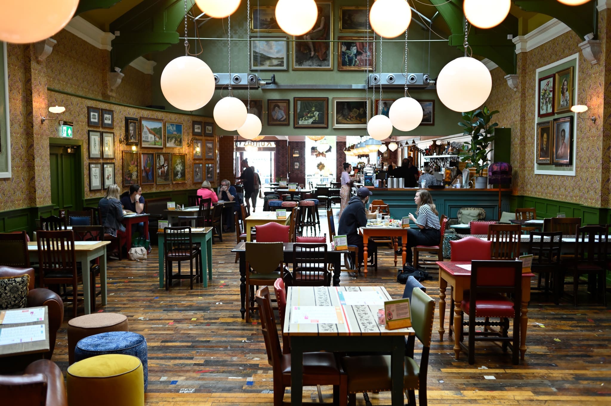 A cozy, eclectic restaurant with wooden floors, colorful mismatched chairs, round hanging lights, and walls decorated with framed art. Several people sit at tables, while the lively Rondo bar is visible in the background.