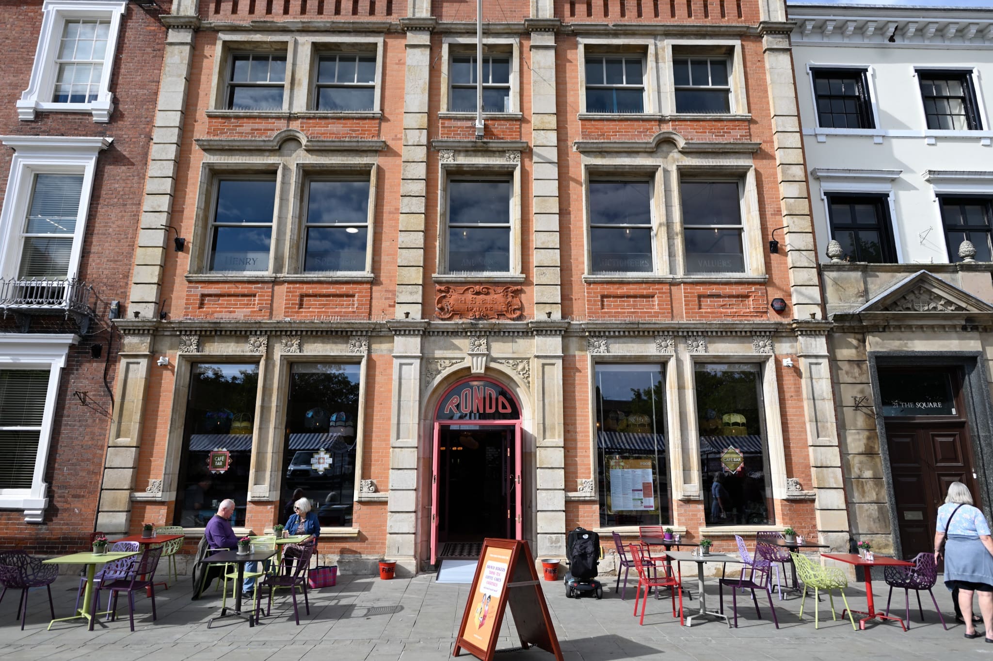 Street view of RIND café with a large arched doorway, outdoor tables and chairs, people sitting and walking, and the tall brick Rondo building featuring large windows and decorative stone detailing.