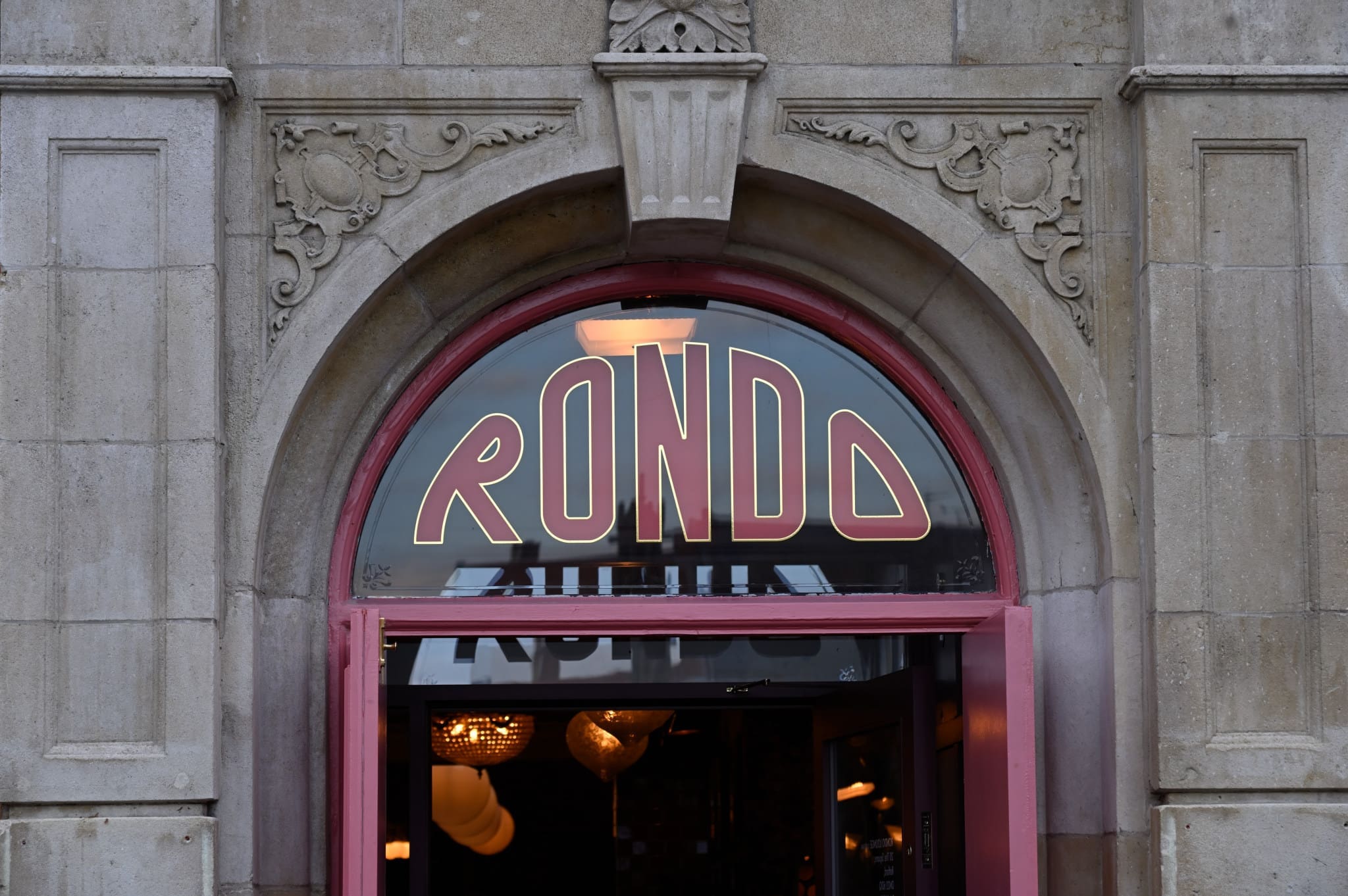 An arched entrance with ornate stone details, featuring a large window above the door displaying "Rondo" in bold, stylized letters. Warm lights and reflections create an inviting atmosphere inside through the glass.