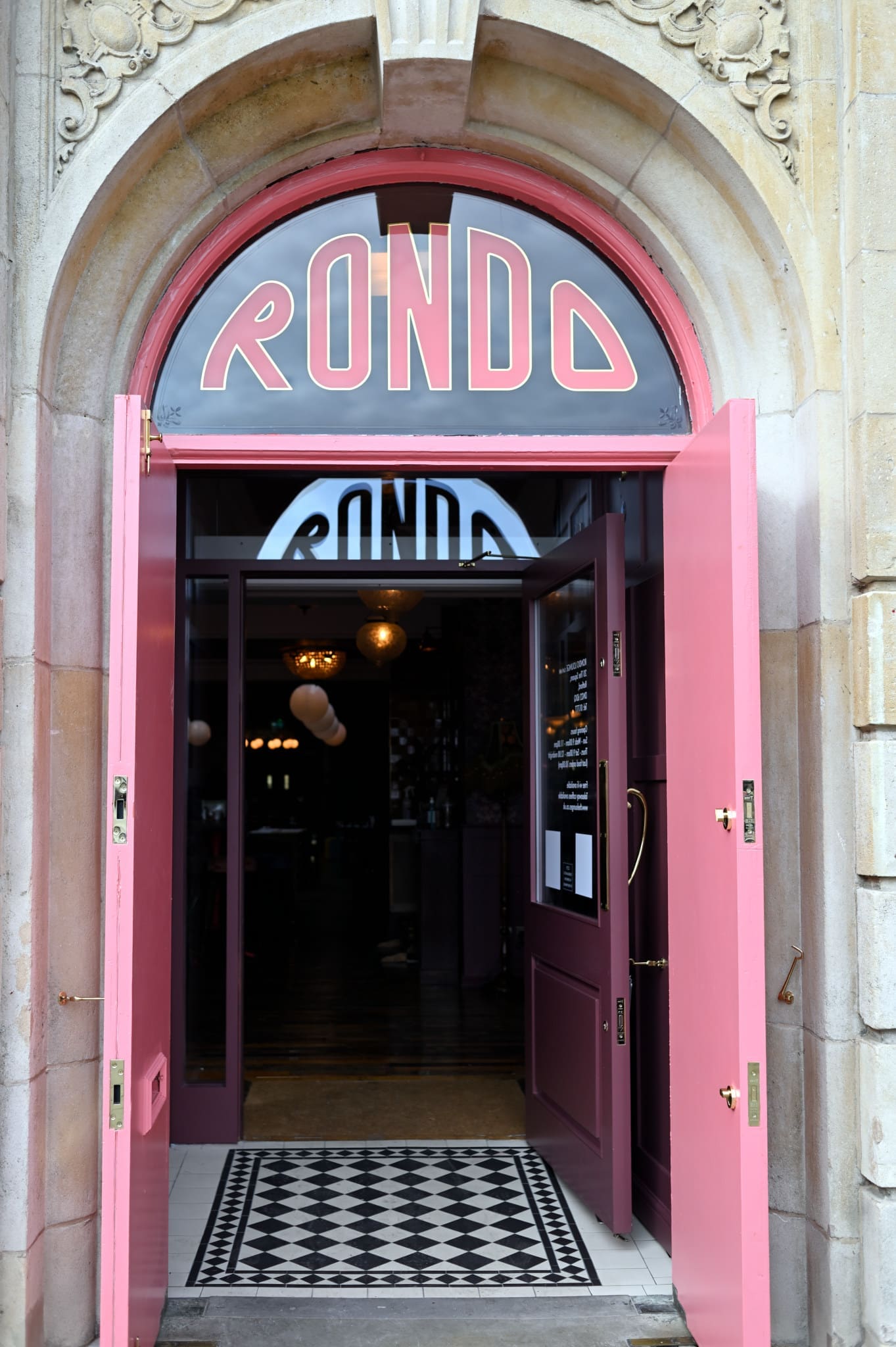 A pink double door stands open, welcoming guests into Rondo, a restaurant or cafe with its name in bold letters above the entrance. The inviting interior features warm lighting and a stylish checkered floor mat.