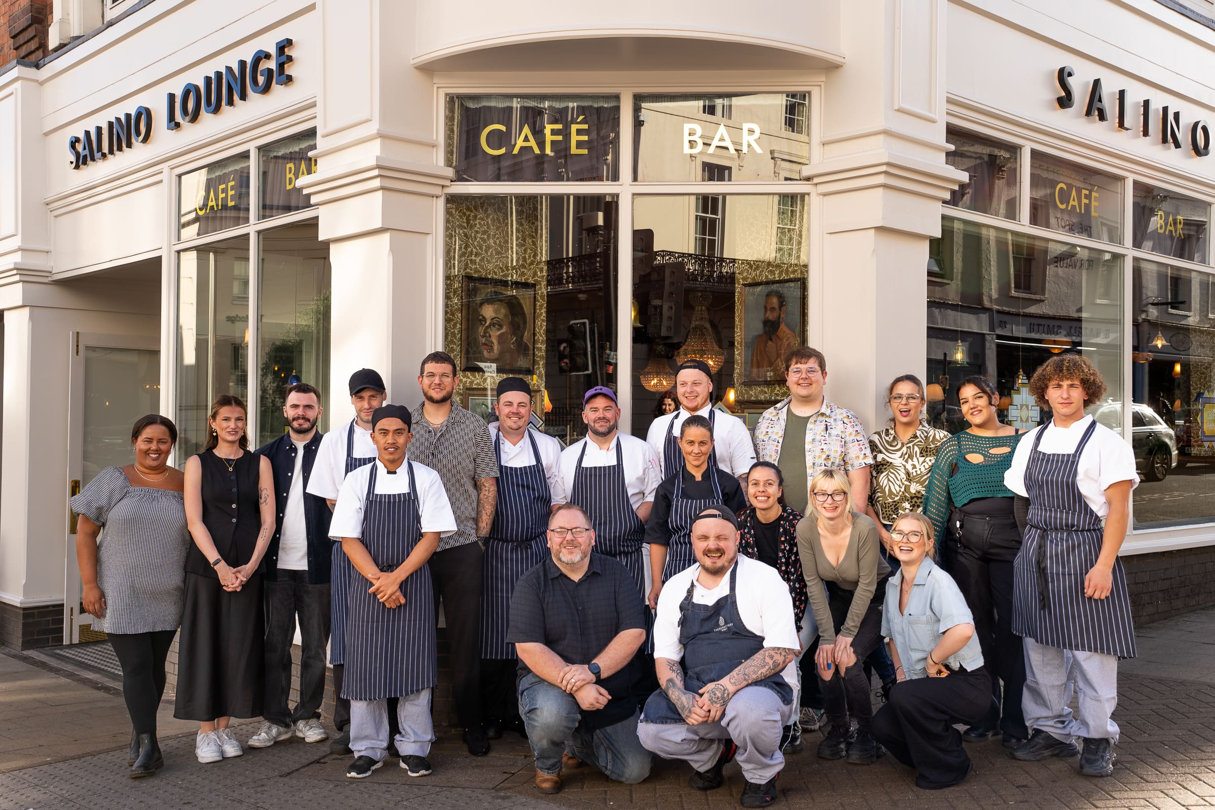 A group of people, including chefs in aprons and staff, pose and smile for a group photo outside the lively Salino Lounge café bar on a city street.