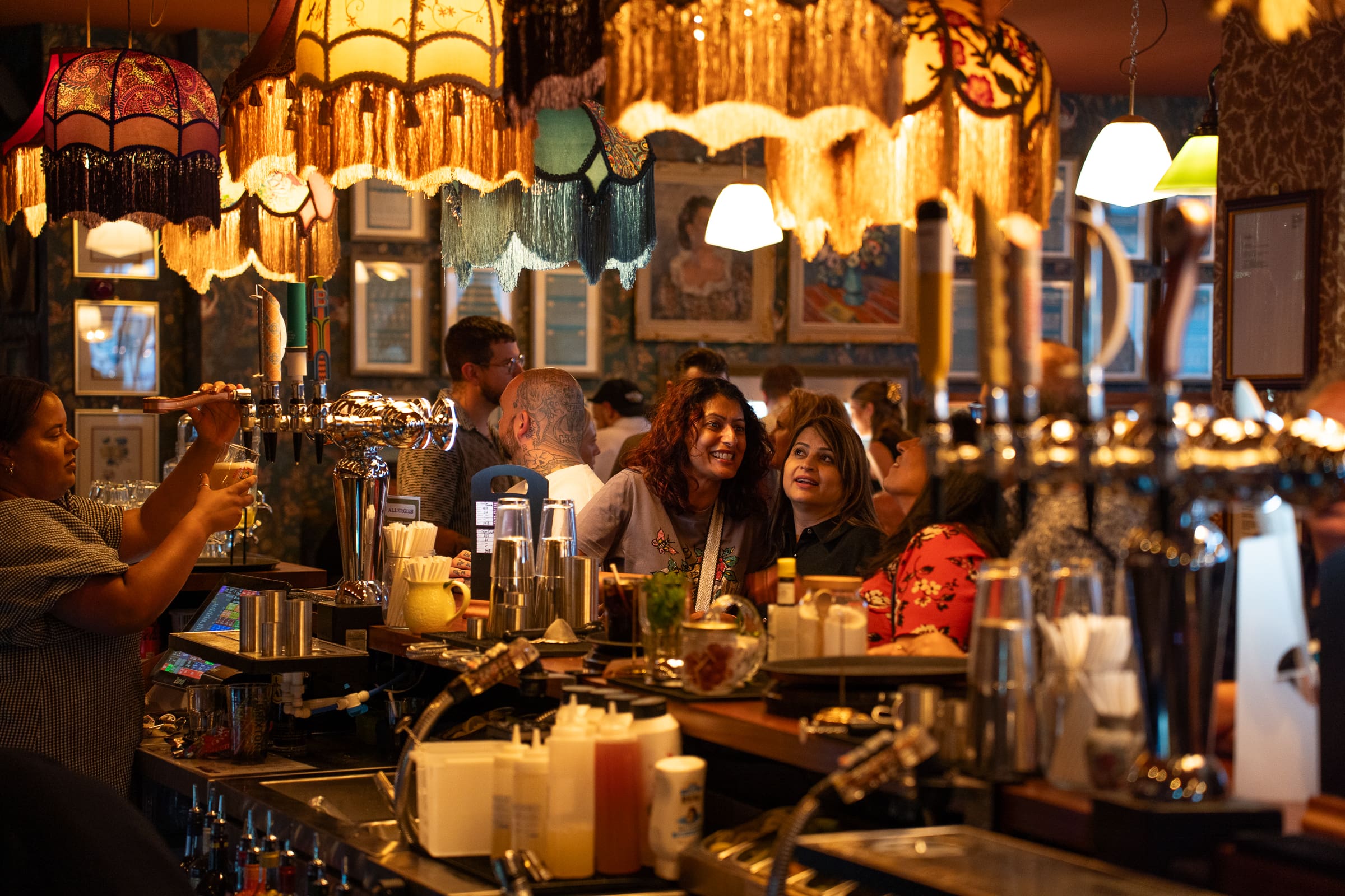 A lively, warmly lit bar like Salino, with vintage lampshades, busy bartenders, and patrons smiling and socializing around the counter. The festive atmosphere is cozy, with drinks and classic bar tools visible on the counter.