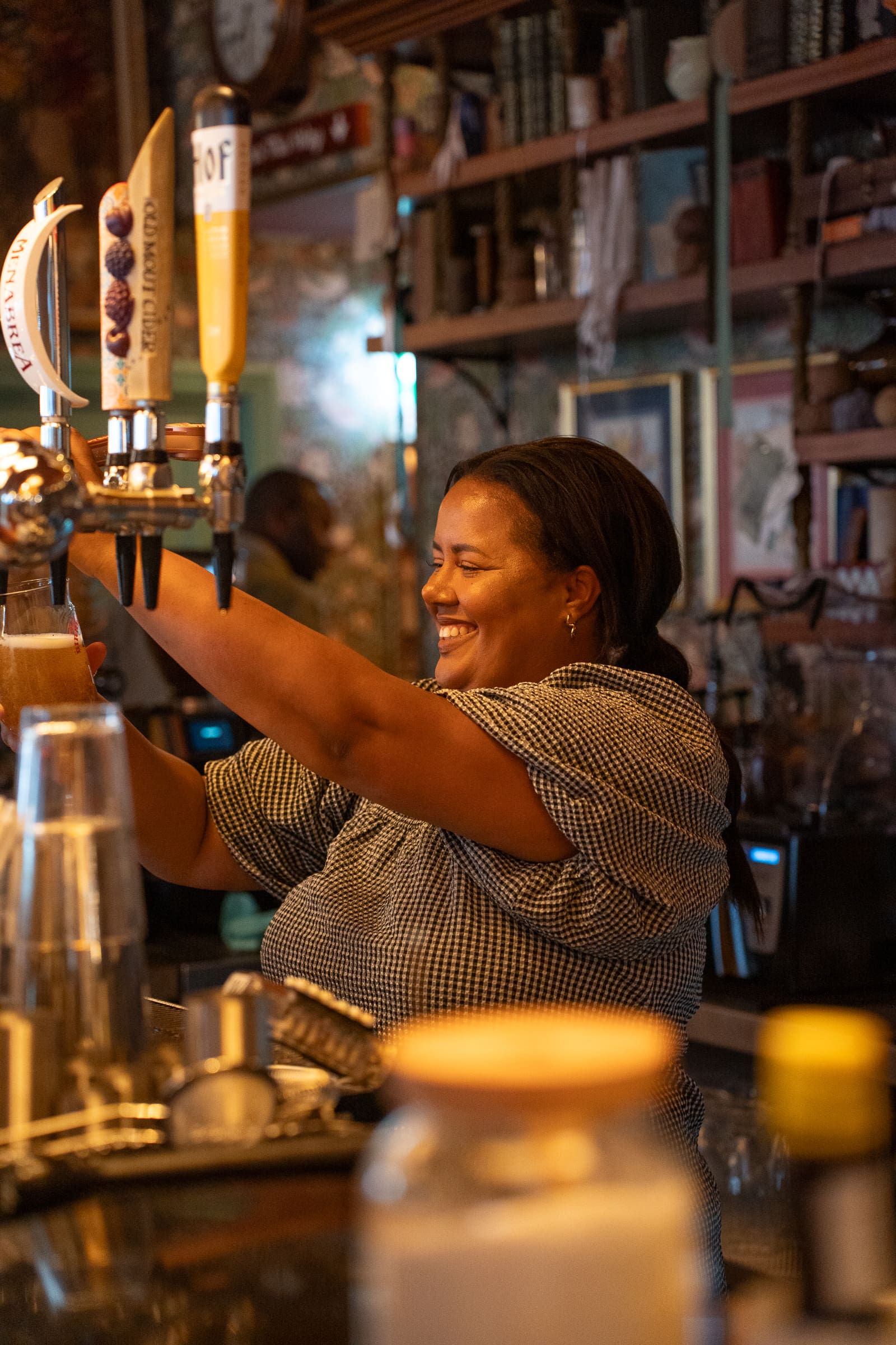 A smiling woman pours a drink from a Salino beer tap behind the bar, surrounded by glasses and shelves filled with various bottles and items. Warm lighting creates a cozy atmosphere.