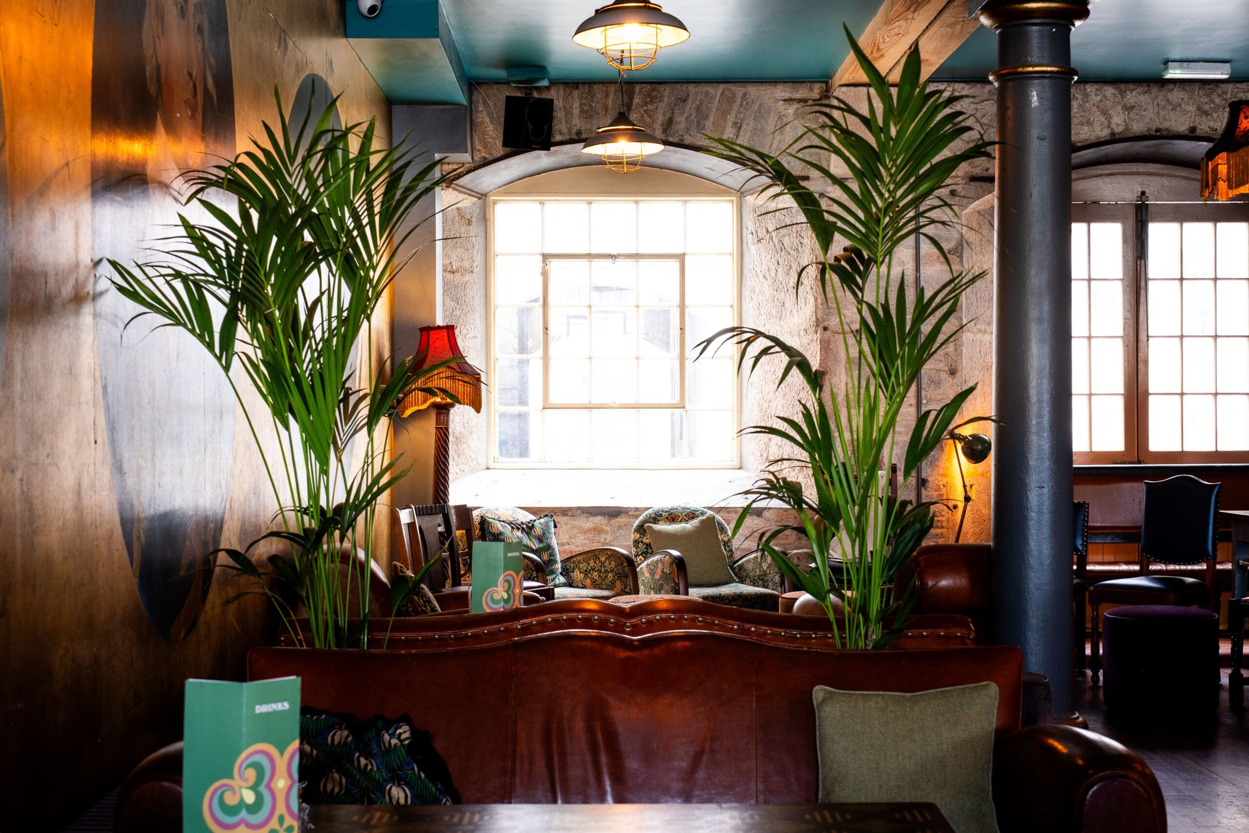 Cozy Seco lounge with leather sofas, tall green plants, and decorative pillows, set near large windows letting in natural light. Warm, eclectic decor includes a red lampshade and wooden accents.