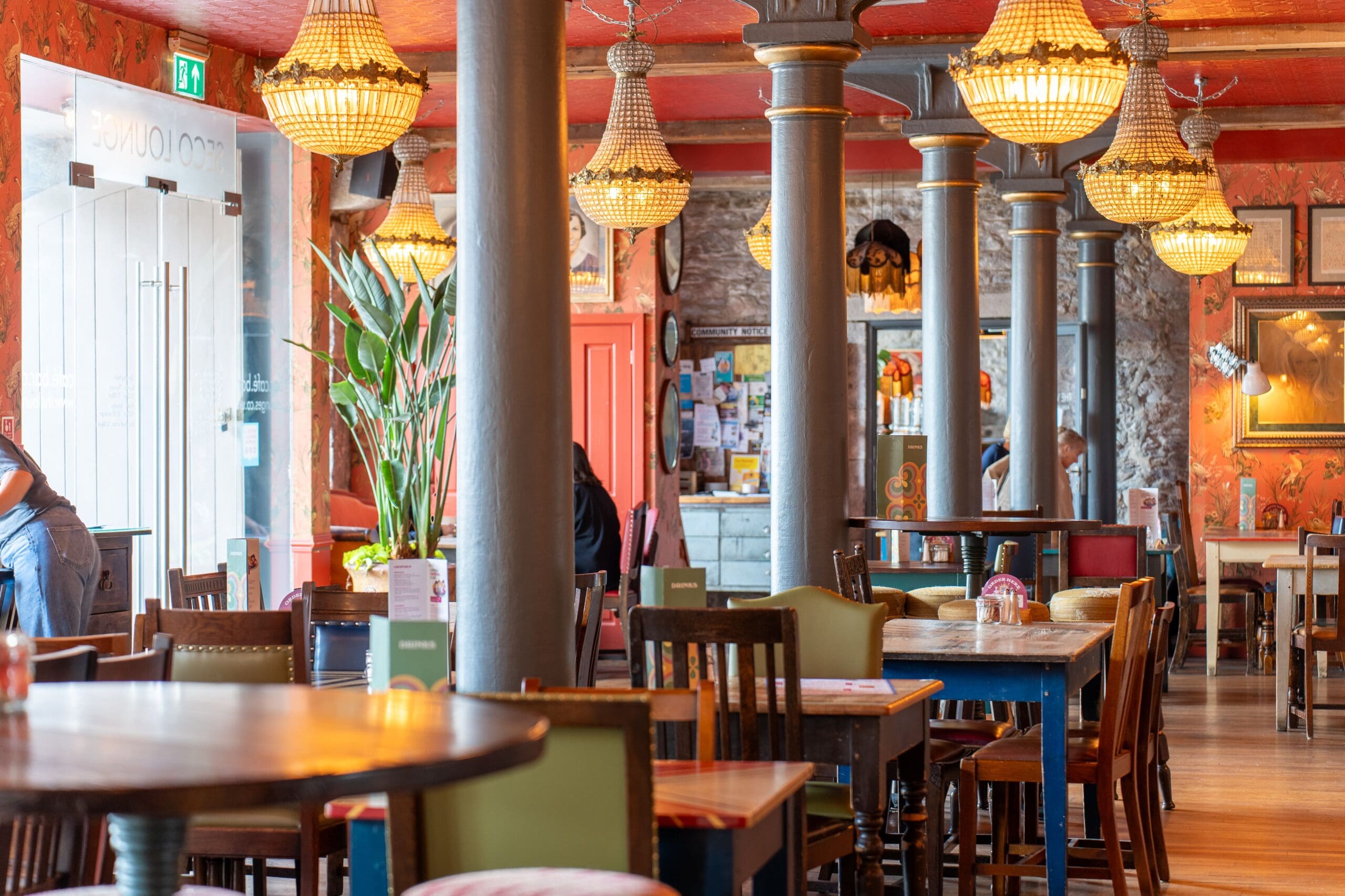 A cozy restaurant interior with colorful walls, chandeliers, tall columns, and eclectic wooden tables and chairs. Warm lighting creates an inviting atmosphere as guests enjoy dishes like Seco; a few people are seated in the background.