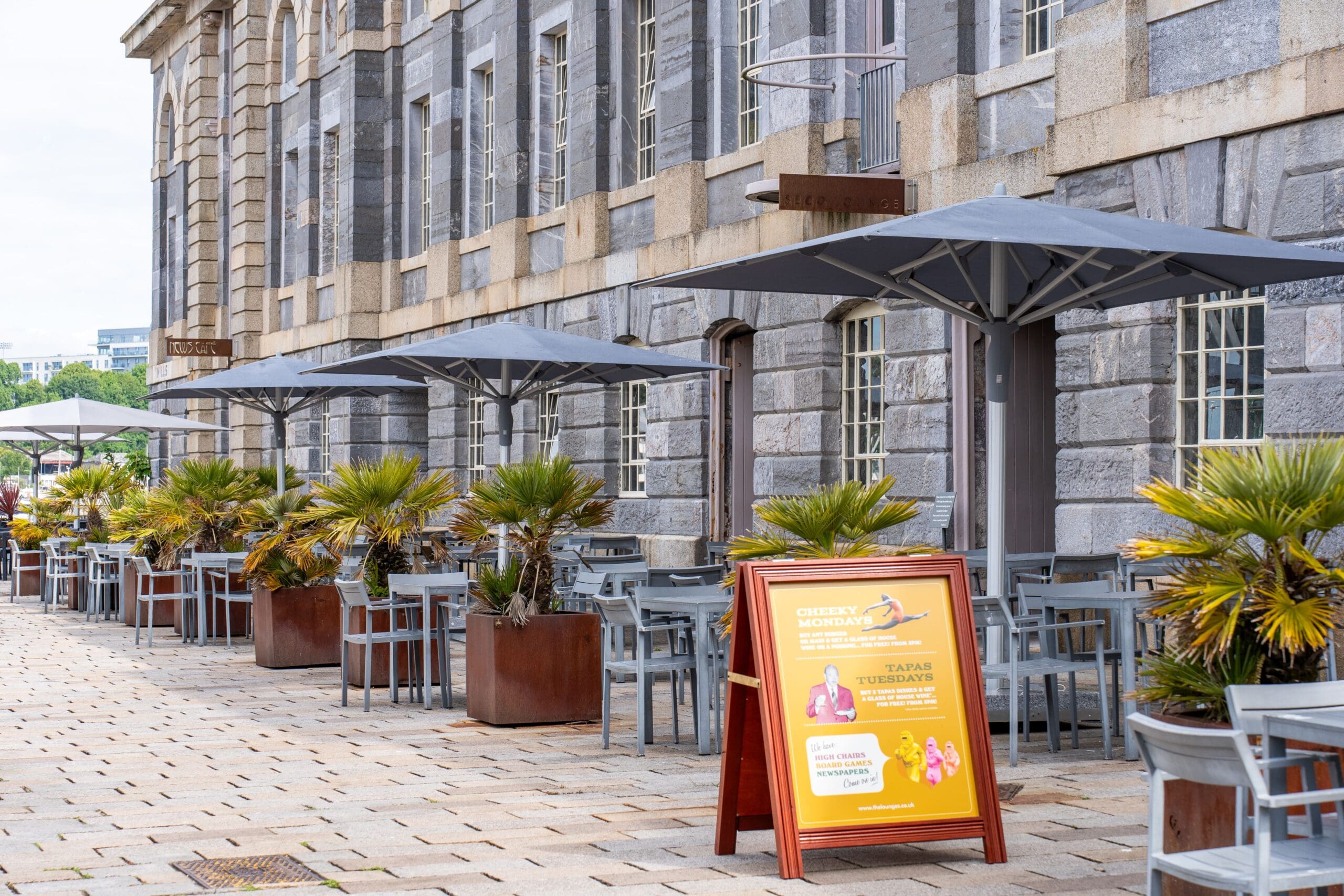 A row of outdoor tables and chairs with umbrellas lines a stone building. Potted palm plants decorate the seating area. A sandwich board advertises tapas, Seco cocktails, and drink specials in front of the restaurant.