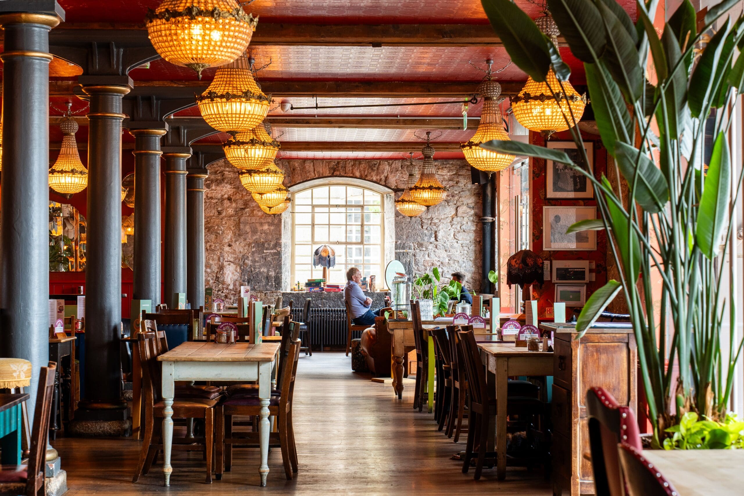 A cozy restaurant interior with wooden tables, mixed chairs, hanging chandeliers, large columns, and plants. Enjoy a plate of seco as sunlight streams through a large window, where a person sits alone by the wall.