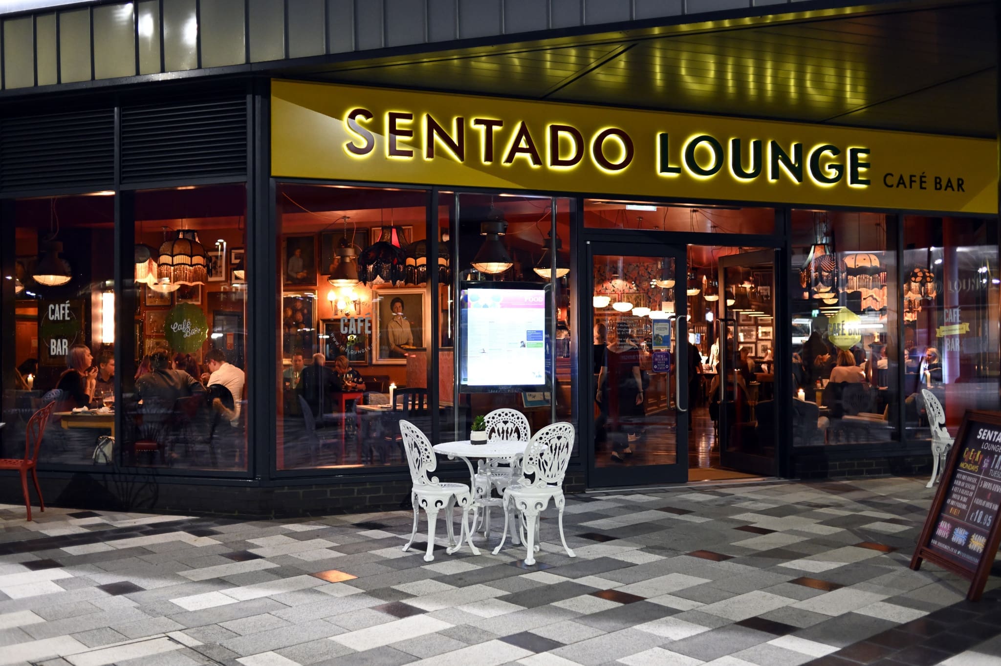 Exterior view of Sentado café bar at night, with warm interior lighting spilling out, people relaxing inside, and two empty white chairs with a table on the tiled pavement outside.