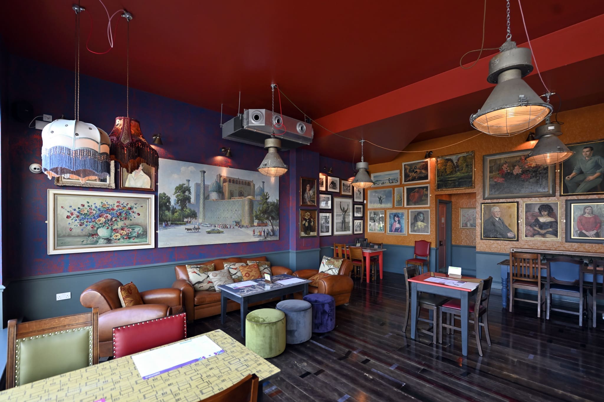 A cozy, stylish café where you can relax sentado amidst eclectic wall art, vintage furniture, colorful stools, wooden tables, hanging pendant lights, and a deep red ceiling—all creating a warm and inviting atmosphere.