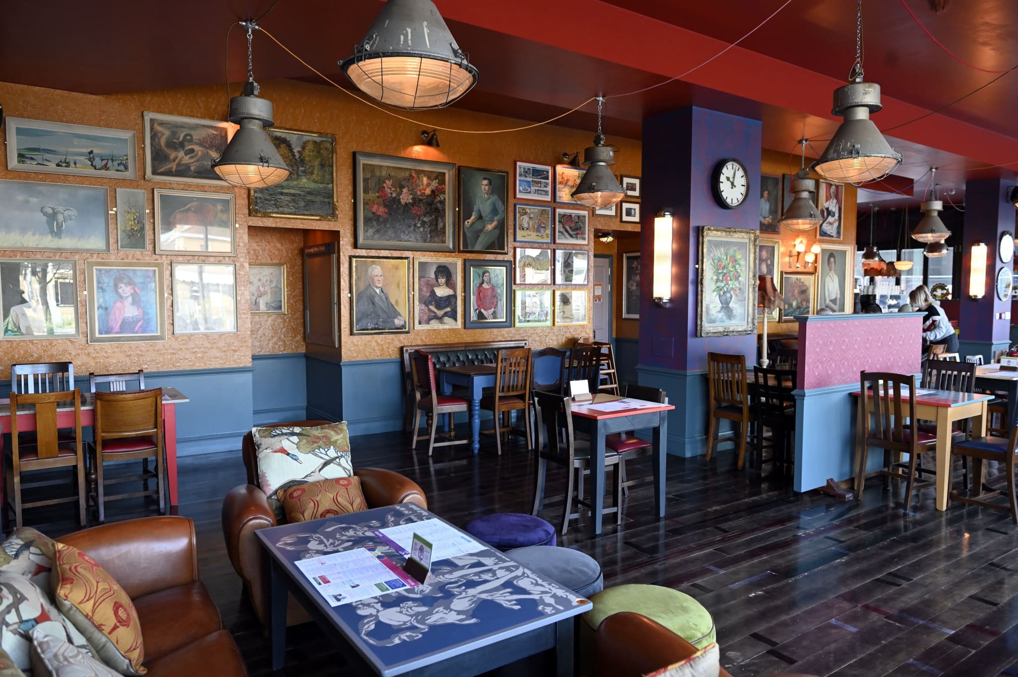 Colorful restaurant interior with wooden tables and chairs, leather armchairs for sentado comfort, and walls decorated with numerous framed artworks. Pendant lights hang from the ceiling, and a clock is mounted on a dark purple column.