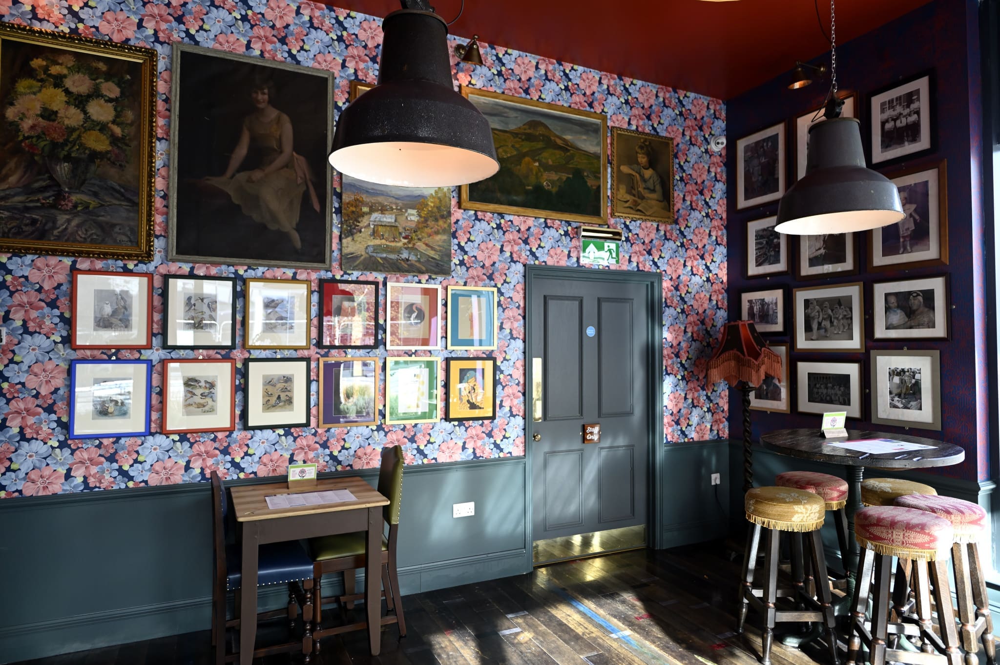 A cozy room with floral wallpaper, framed art and photos on the walls, a small table with two chairs for sentado moments, and a corner with three stools around a small round table, lit by two large hanging lamps.