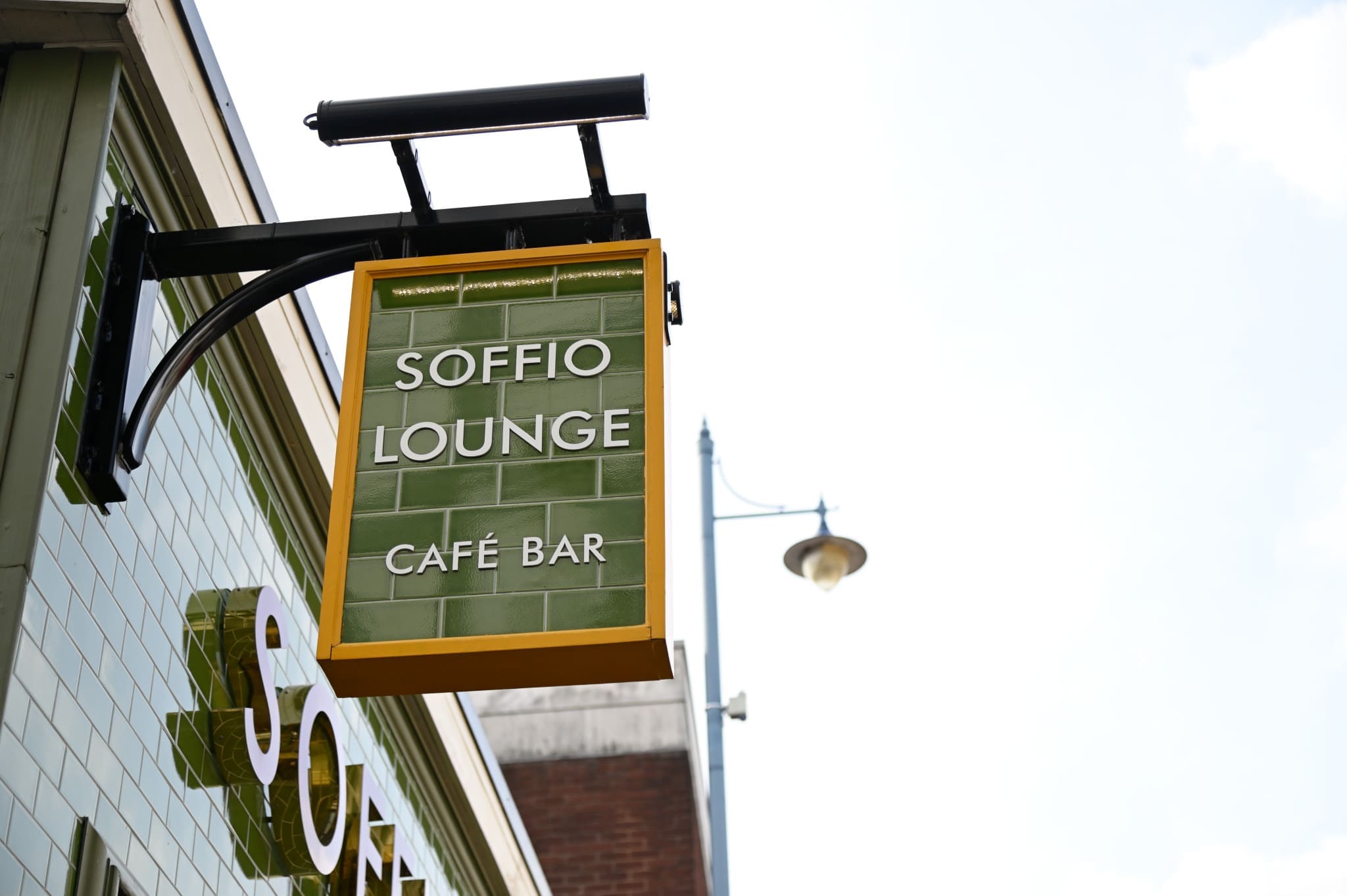 A green and yellow sign for Soffio Lounge Café Bar hangs outside a building, displaying "Soffio" in white letters, with a streetlamp and clear sky in the background.