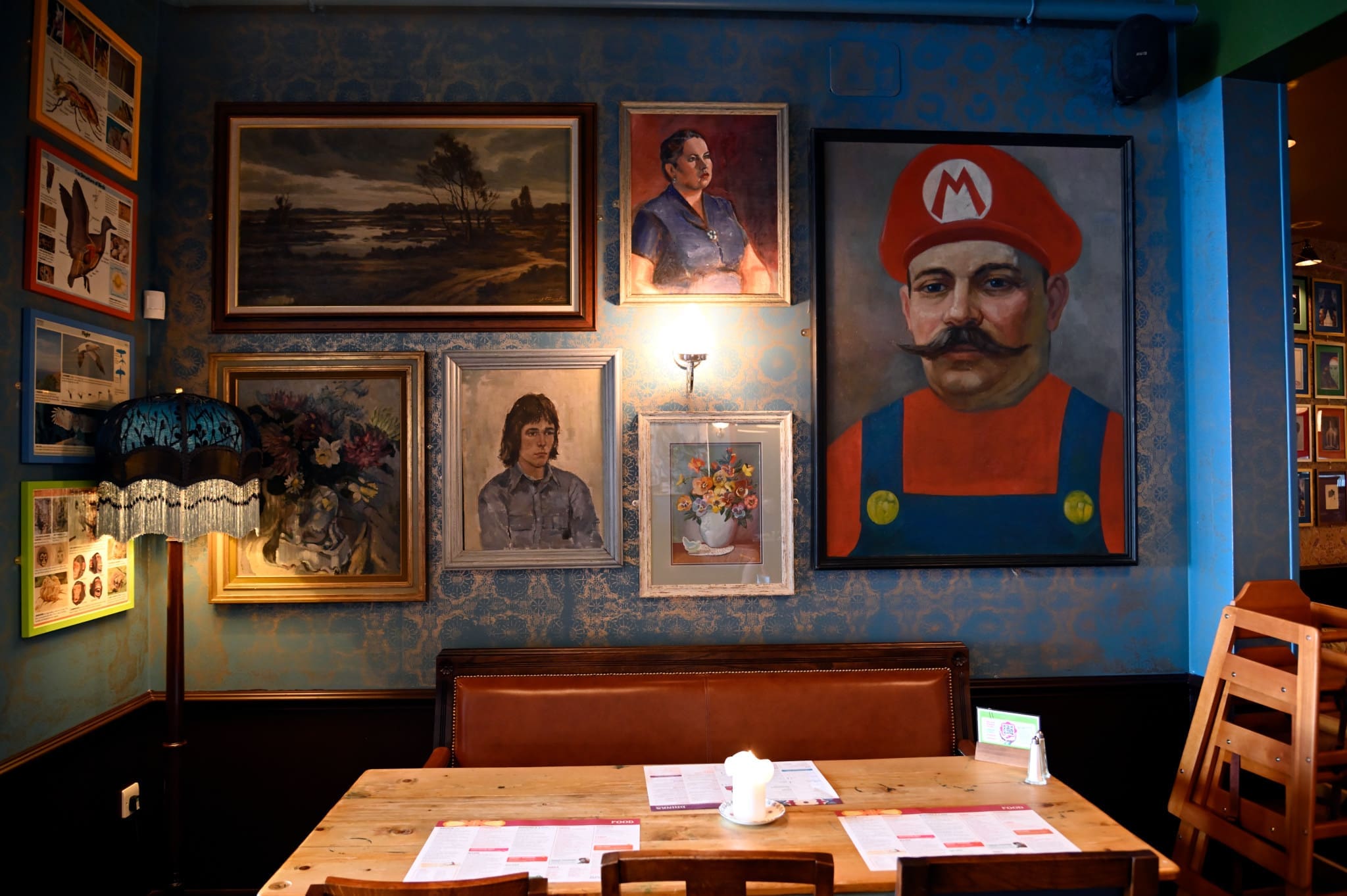 A cozy Soffio restaurant interior with a wooden table and chairs, vintage lamp, and a wall decorated with eclectic framed paintings, including a large portrait of a man in a Mario costume. Soffio menus are placed on the table.