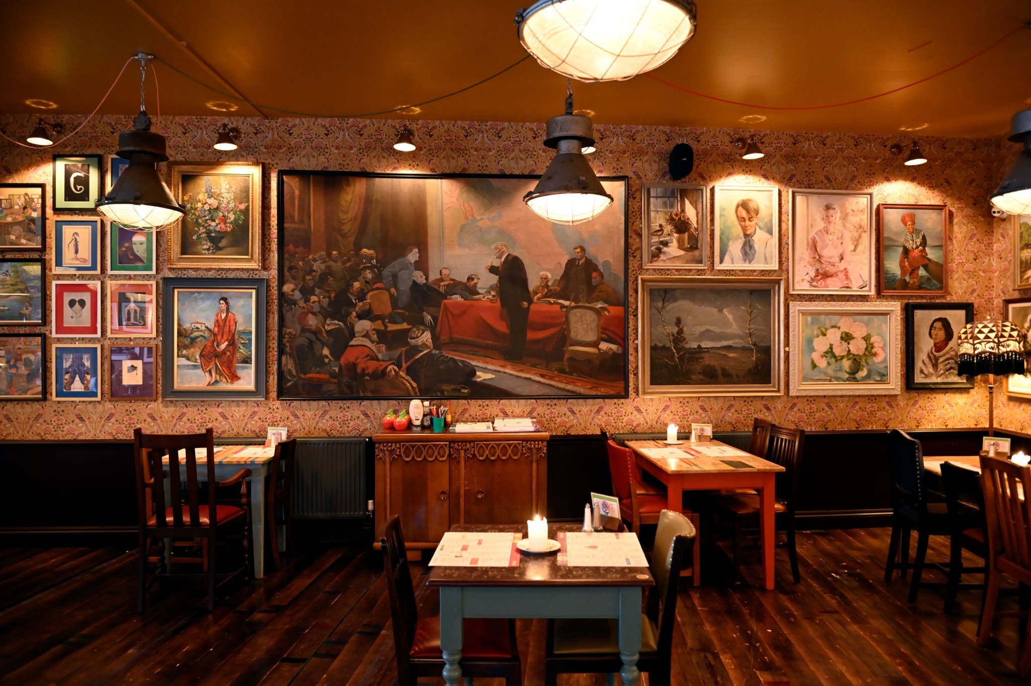 A cozy restaurant interior with wooden floors, empty tables, and chairs. The walls are decorated with numerous framed paintings and portraits, gently illuminated by warm Soffio overhead lights.