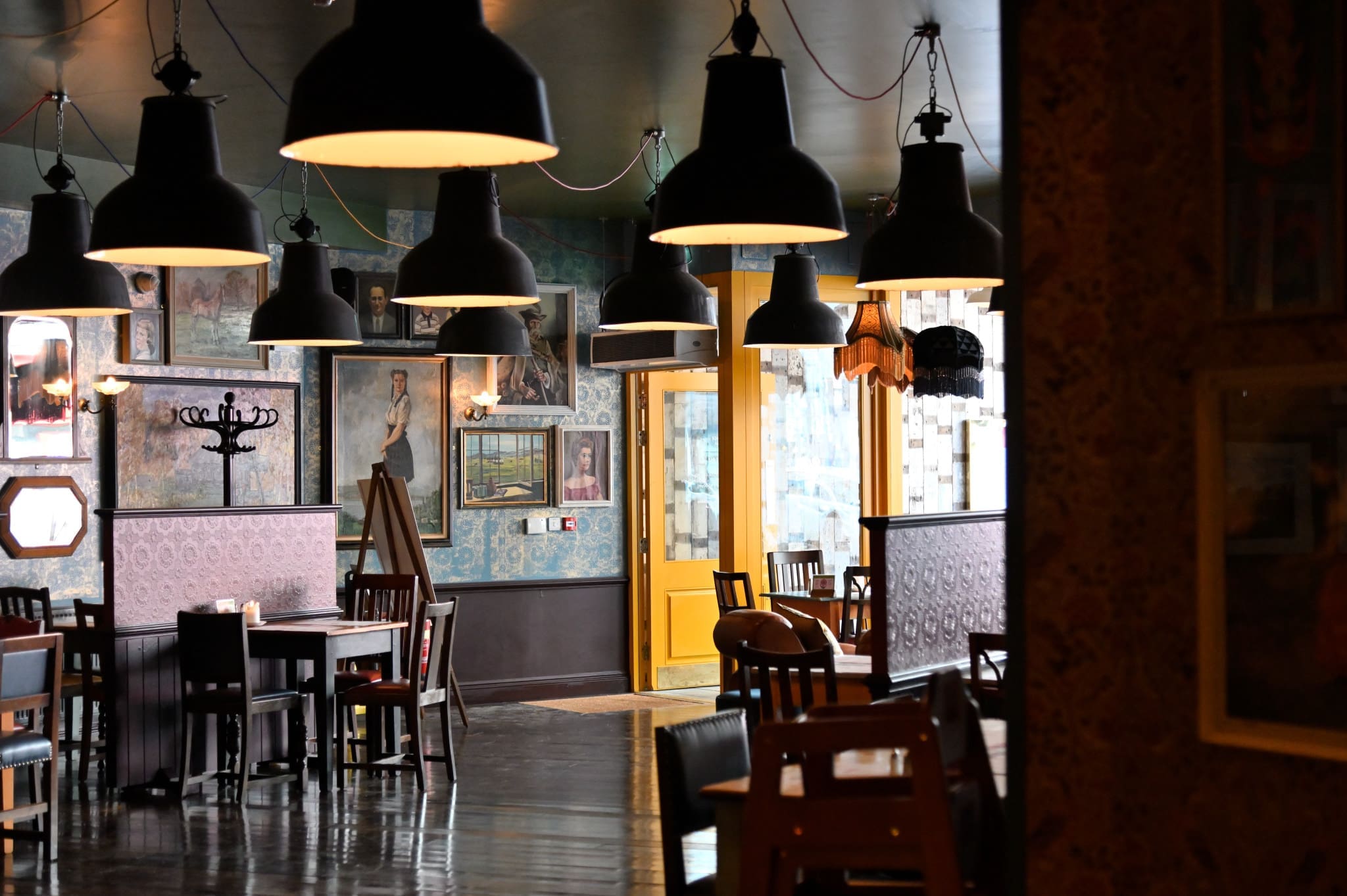 Cozy café interior with Soffio dark hanging lamps, wooden tables and chairs, paintings on wallpapered walls, and a bright yellow door leading to another room. The softly lit Soffio space feels inviting and warm.