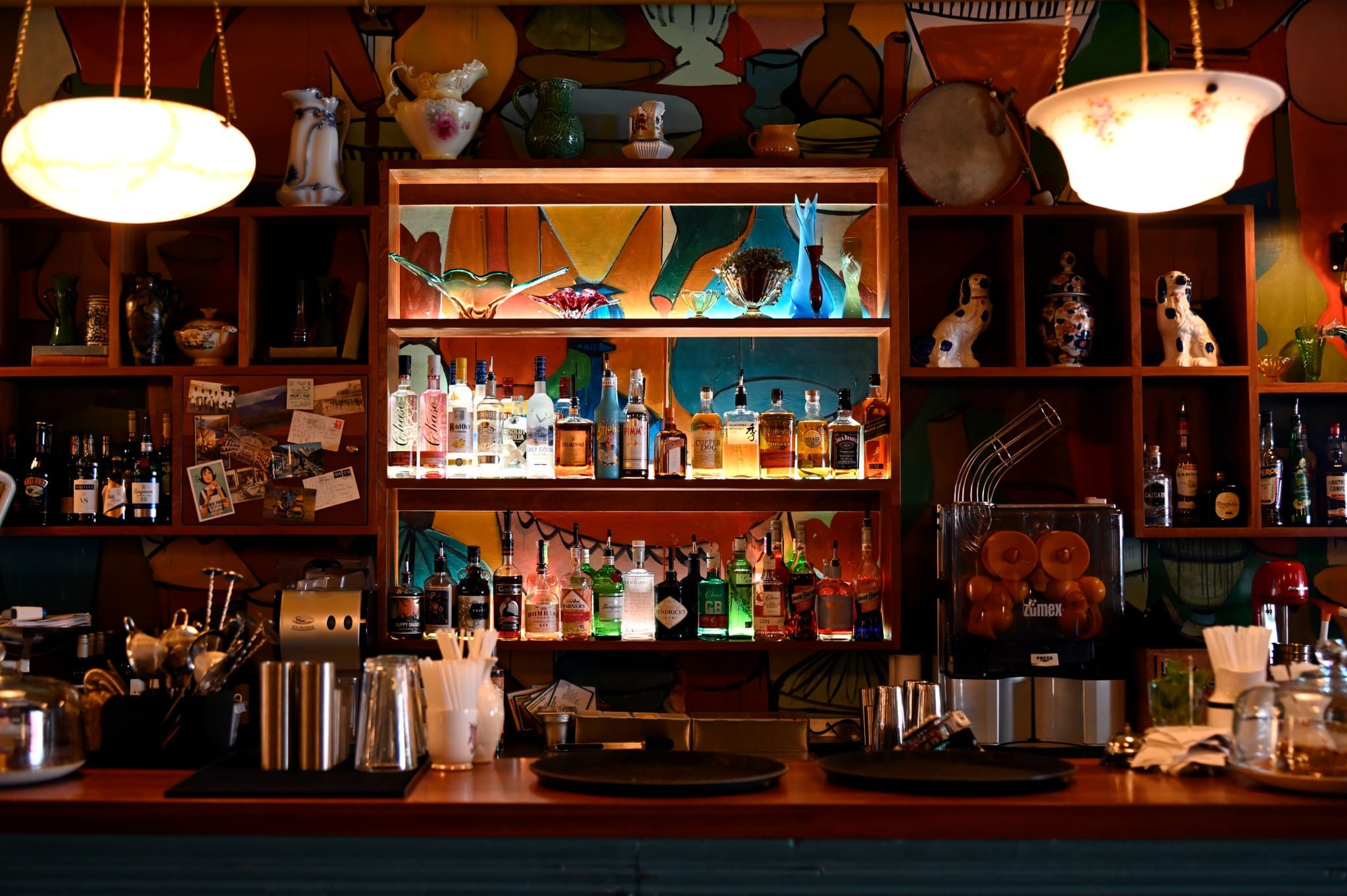 A colorful, eclectic bar by Soffio features various bottles of alcohol on shelves, surrounded by decorative vases, ceramic dogs, lamps, and an orange juicer, all set against a vibrant mural background.