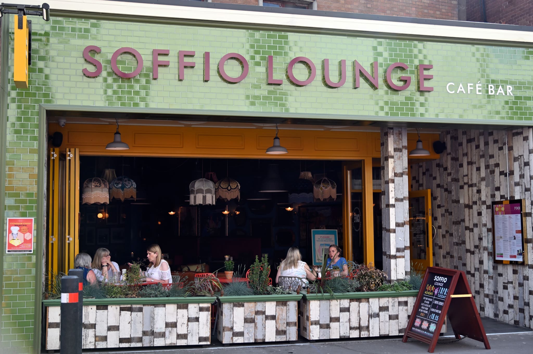 Four women sit at a table outside Soffio Lounge Café Bar, which features green tiles, large windows, and plants along the front. The name "Soffio" is displayed in large letters above the entrance.