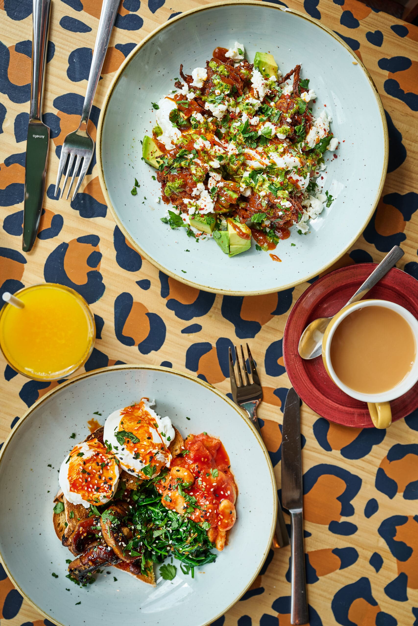 Two plates of brunch food with eggs, vegetables, and herbs sit on a leopard-print table alongside a glass of orange juice and a cup of coffee with milk. Cutlery is placed next to each plate.