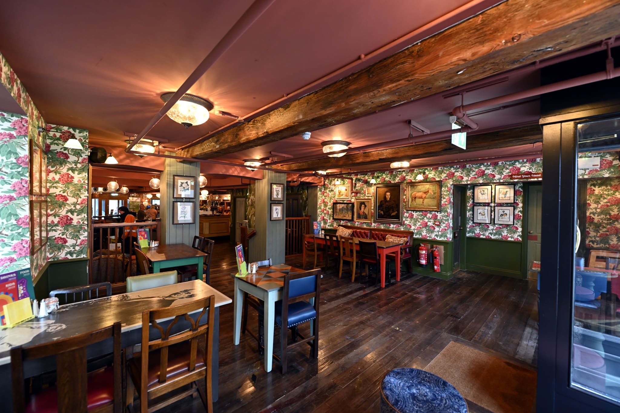 A cozy tambo-inspired pub interior with floral wallpaper, wooden beams, and a mix of wooden tables and chairs. Warm lighting and framed pictures decorate the walls, creating a welcoming atmosphere.