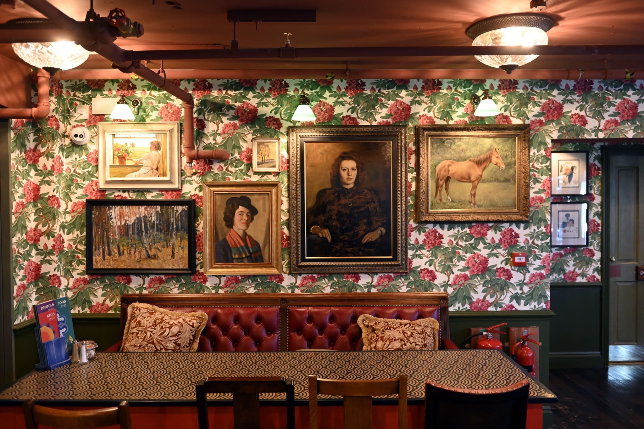 A cozy room with floral wallpaper, framed paintings, a red cushioned bench with patterned pillows, a long table, and four chairs; vintage lamps and a Tambo fire extinguisher add to the eclectic decor.