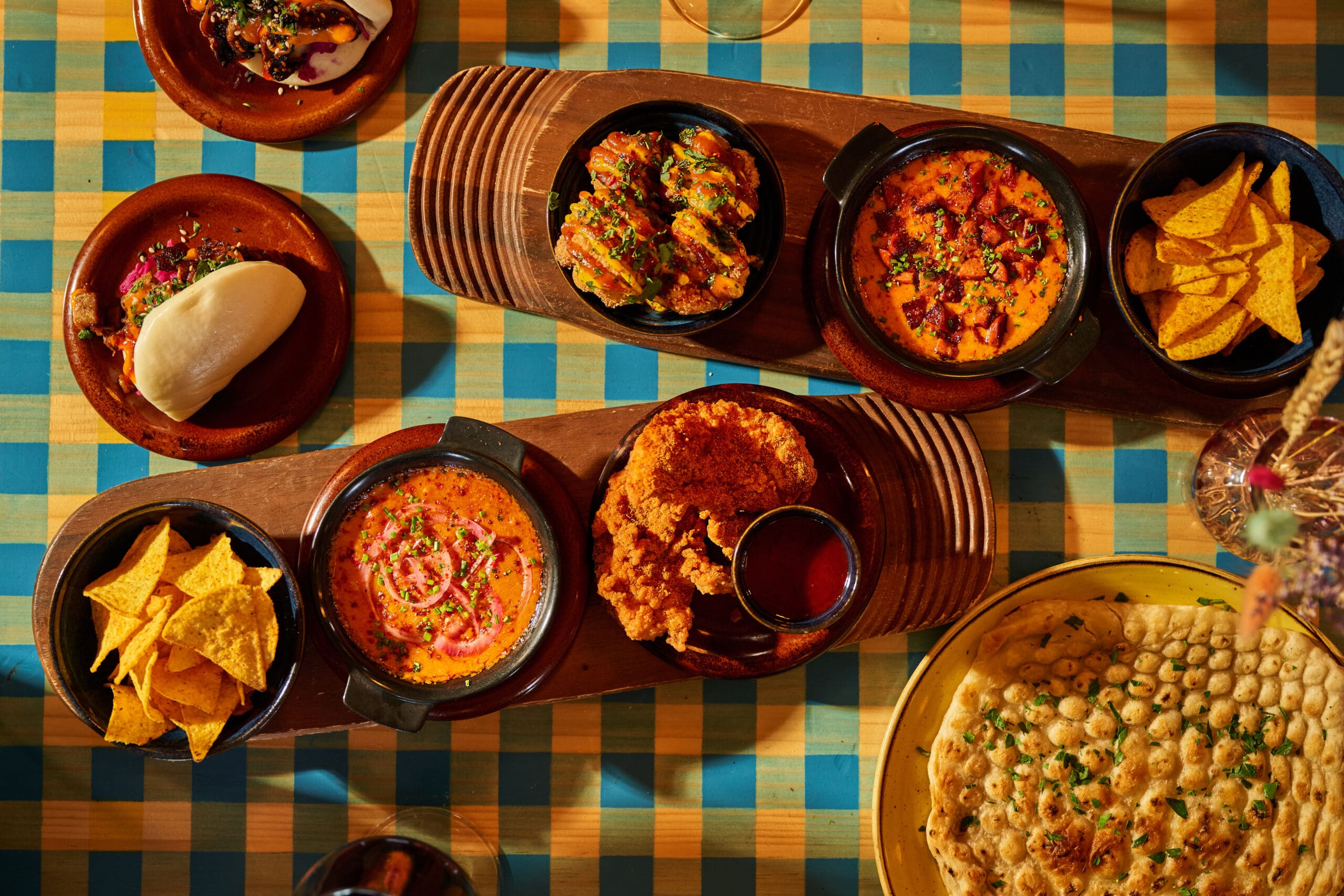 An overhead view of a table with various dishes, including curry, naan, tortilla chips, fried chicken, bao, and appetizers, all arranged on wooden boards and plates over a blue and yellow checkered tablecloth.