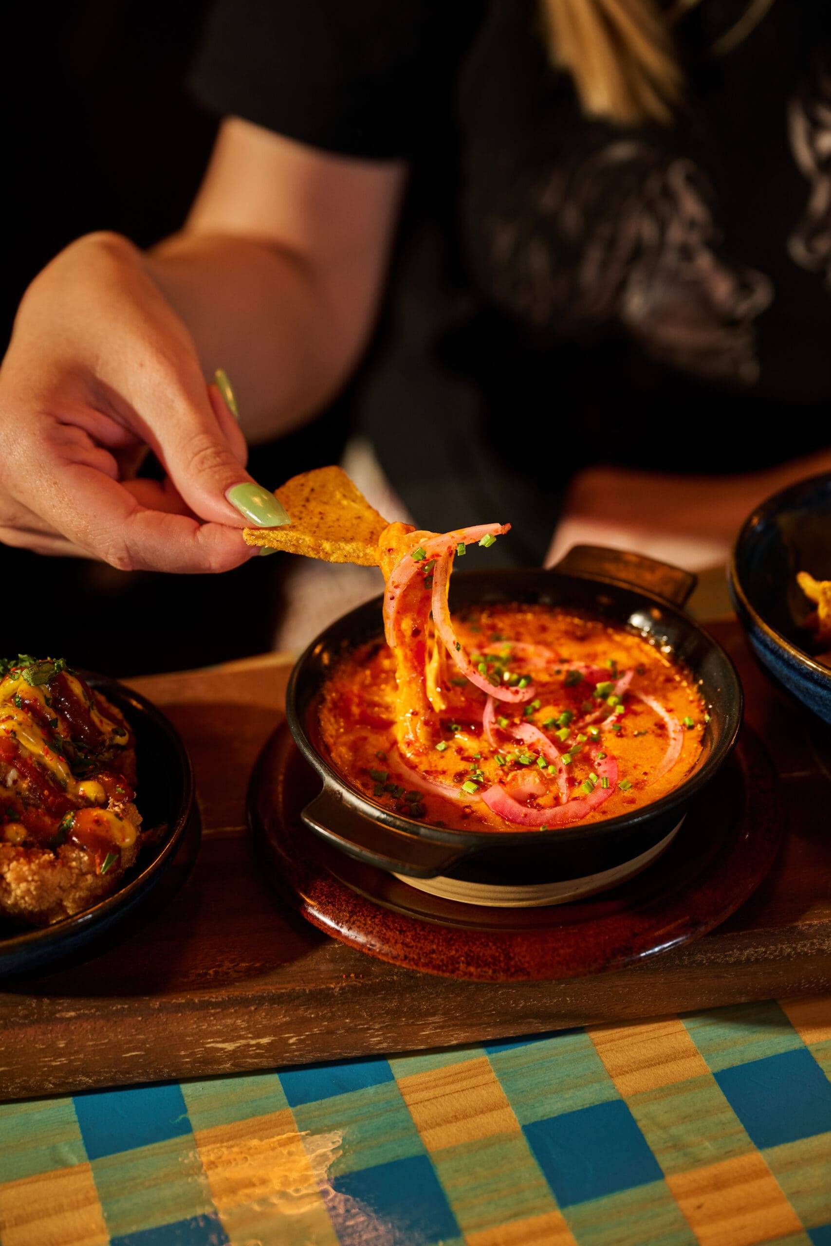 A person dips a tortilla chip into a bowl of creamy, orange-colored dip garnished with herbs and red onions, with other dishes and a checkered table visible on the side.